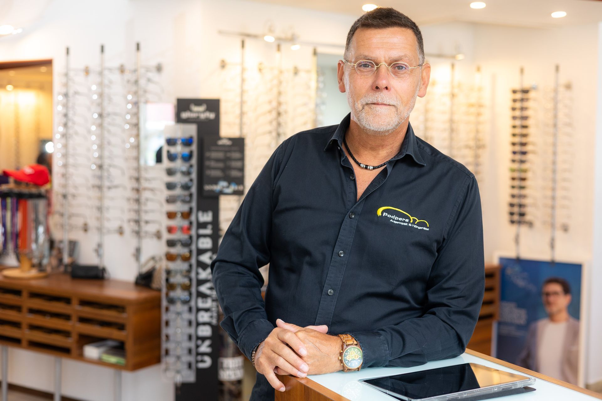 Ein Mann mit Brille steht an einer Theke in einem Brillengeschäft mit Brillendisplays und Holzbeschlägen.