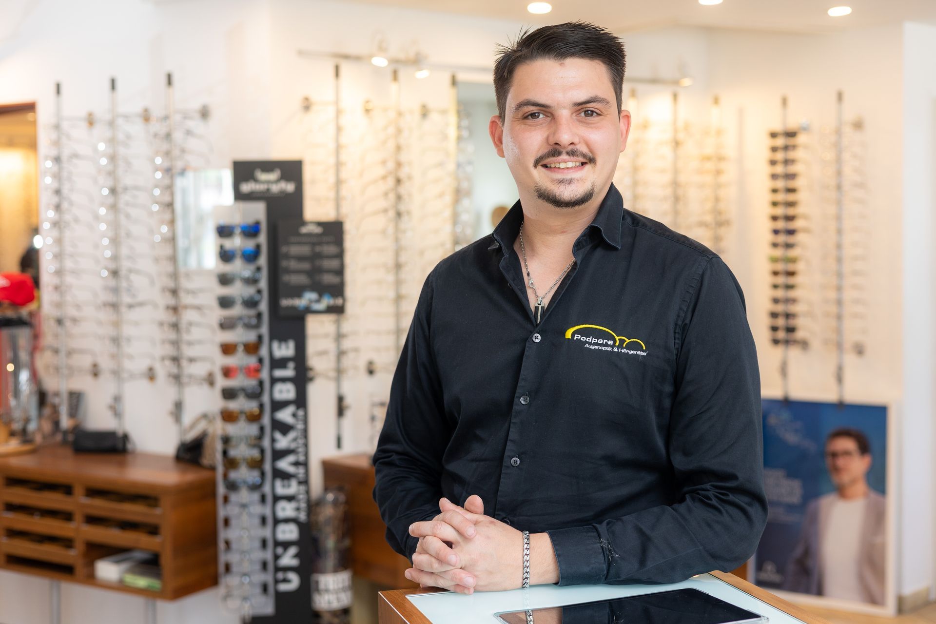 Lächelnder Mann im Brillengeschäft, schwarzes Hemd mit Logo, umgeben von Brillengestellen.