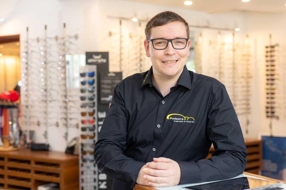 Lächelnder Mann mit Brille in einem Optikergeschäft, schwarzes Hemd, umgeben von Brillengestellen.