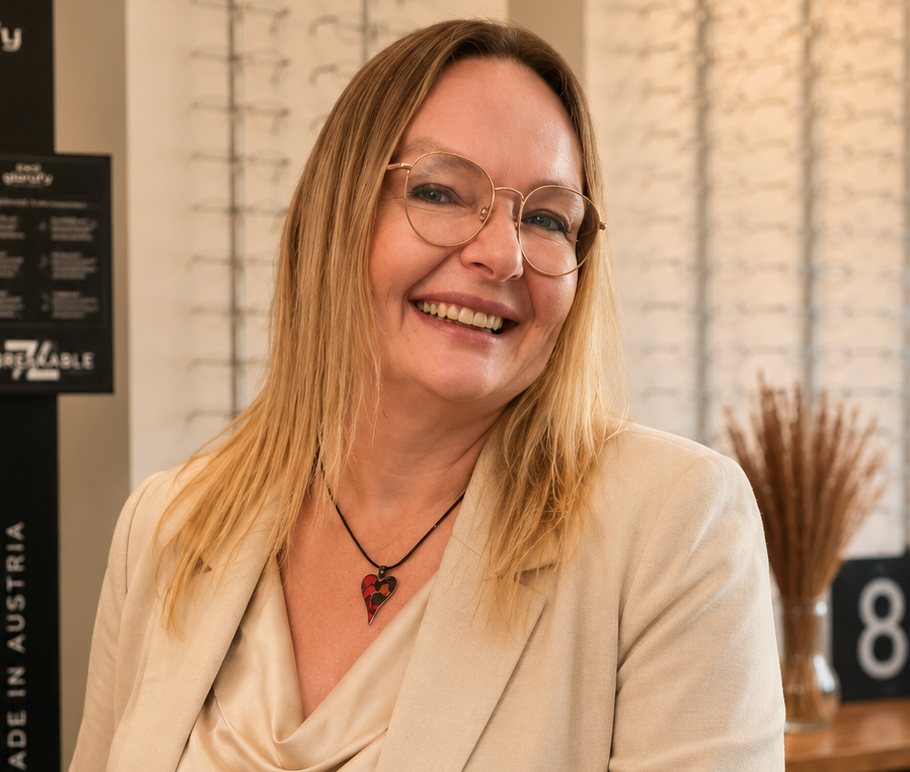Eine lächelnde Frau mit Brille in einer beigefarbenen Bluse steht in einem schwach beleuchteten Laden- oder Galerieinterieur.