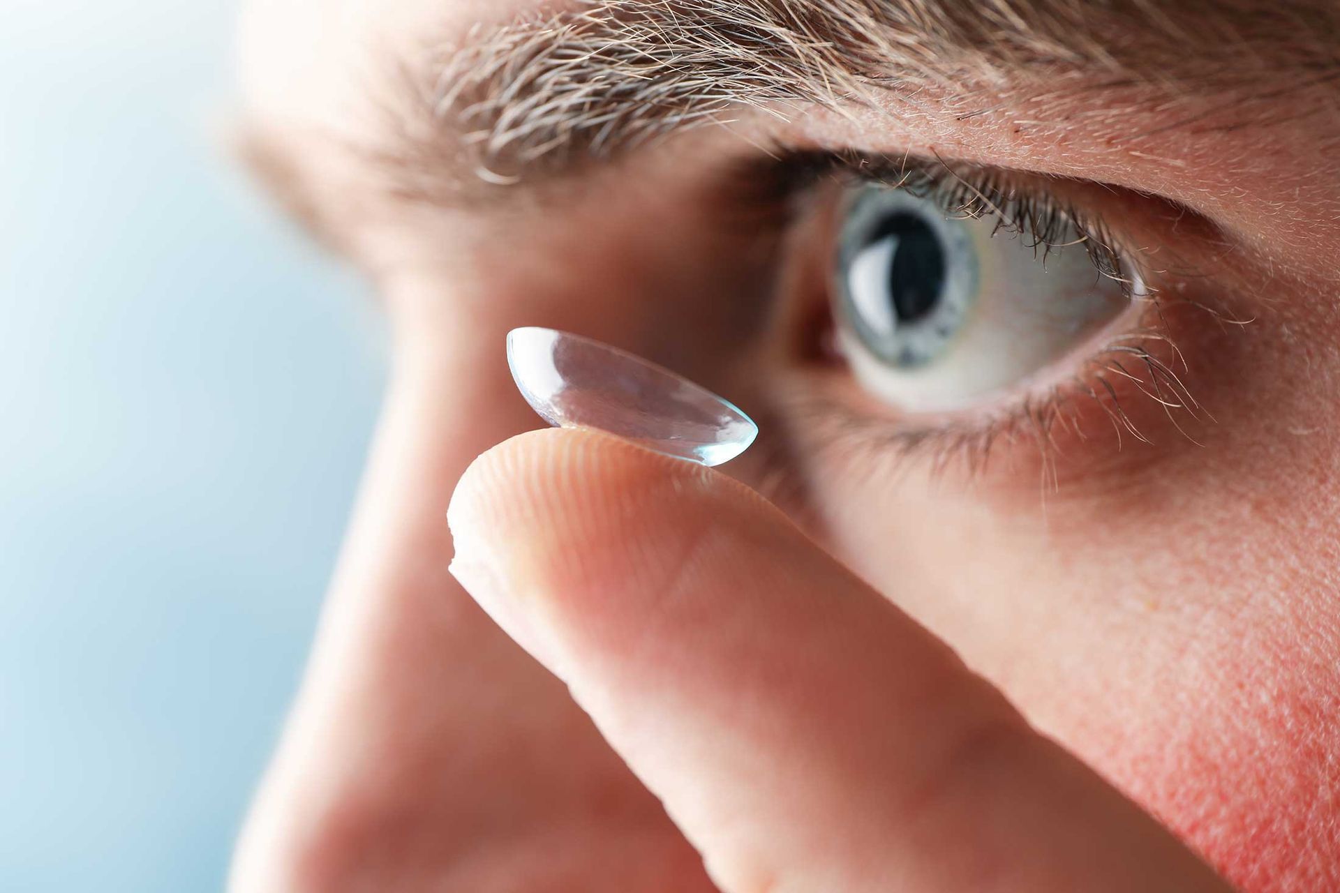 Person hält Kontaktlinse ins Auge und bereitet sich darauf vor, sie einzusetzen. Blaues Auge im Fokus.