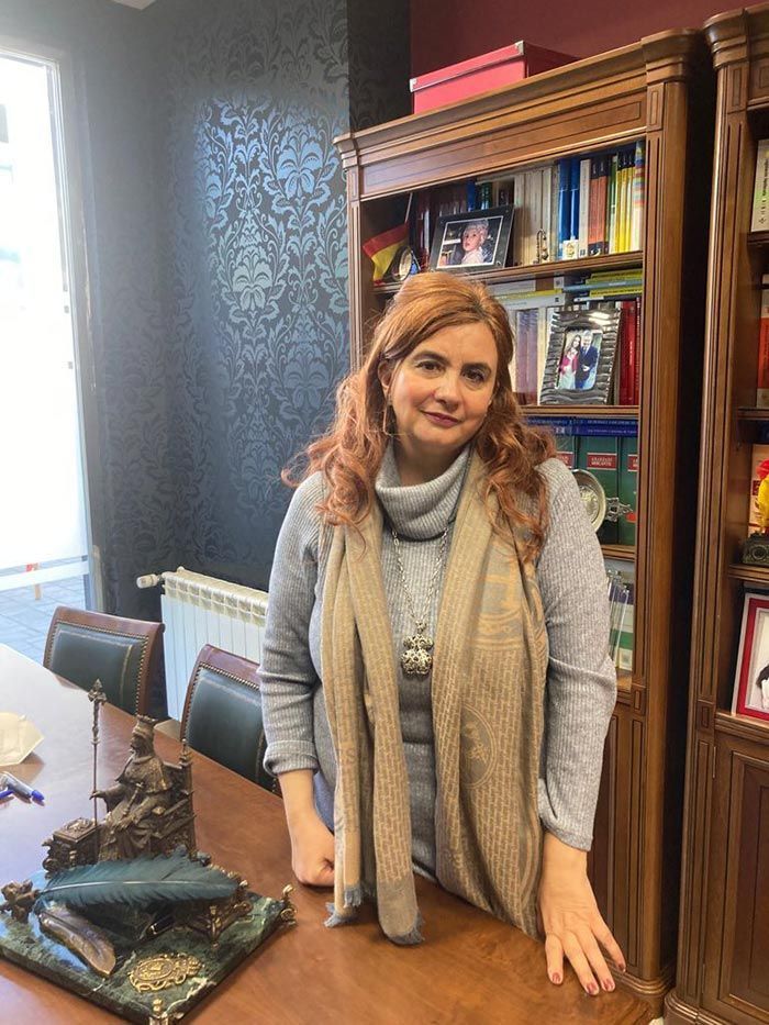 Una mujer con un suéter gris y una bufanda se encuentra en una oficina con una estantería.