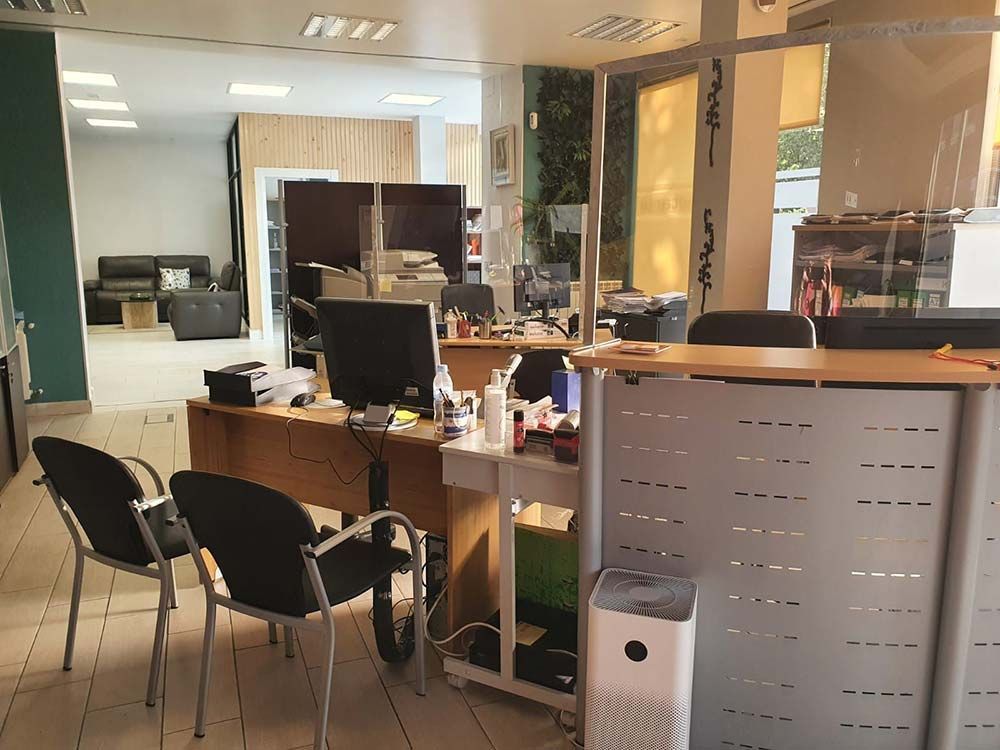 Área de recepción con escritorios, computadoras, sillas y mamparas protectoras. Paredes blancas, detalles en madera y purificador de aire.