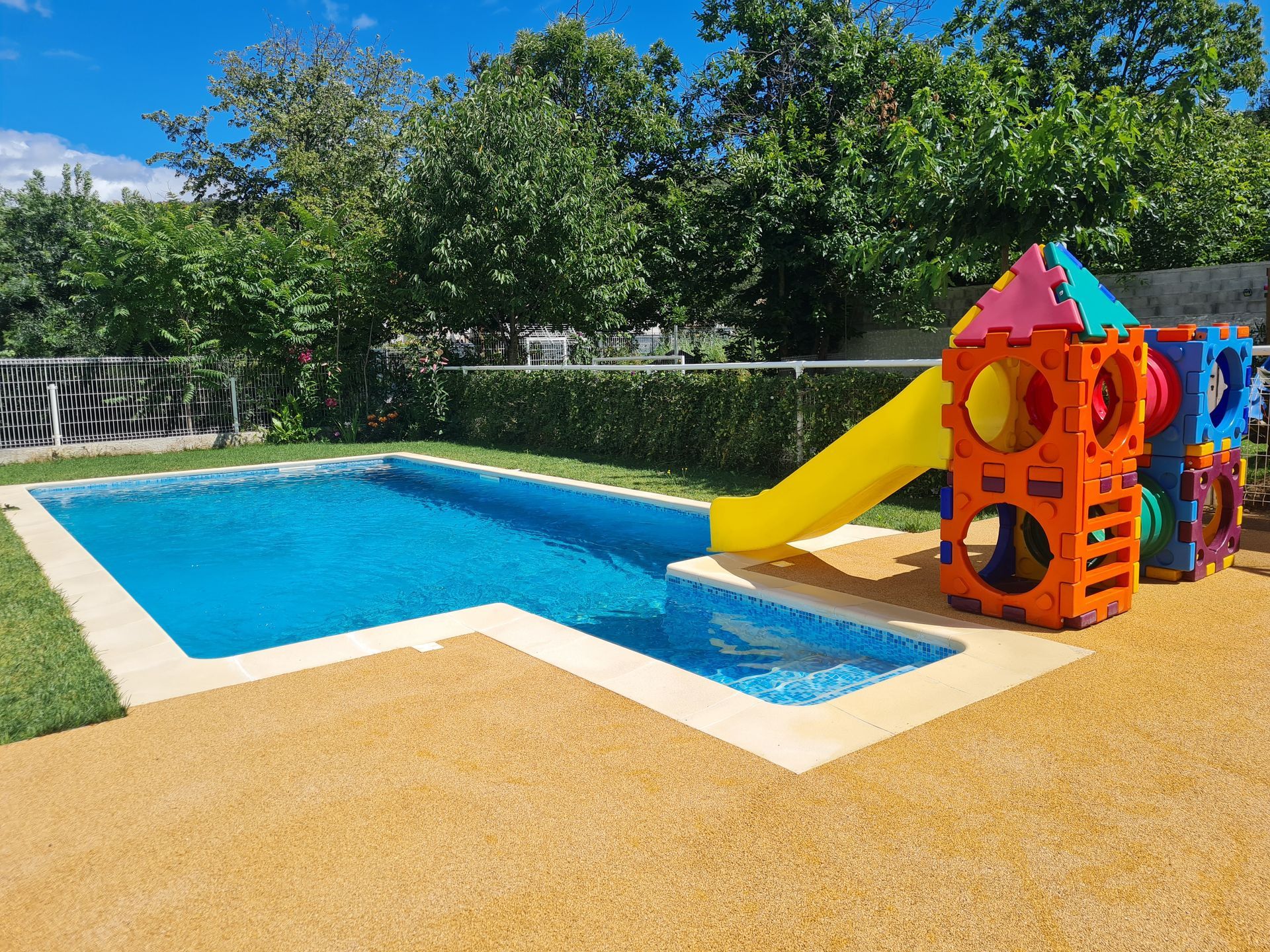 Piscine avec plage en résine beige et aire de jeux pour enfants dans un jardin arboré.