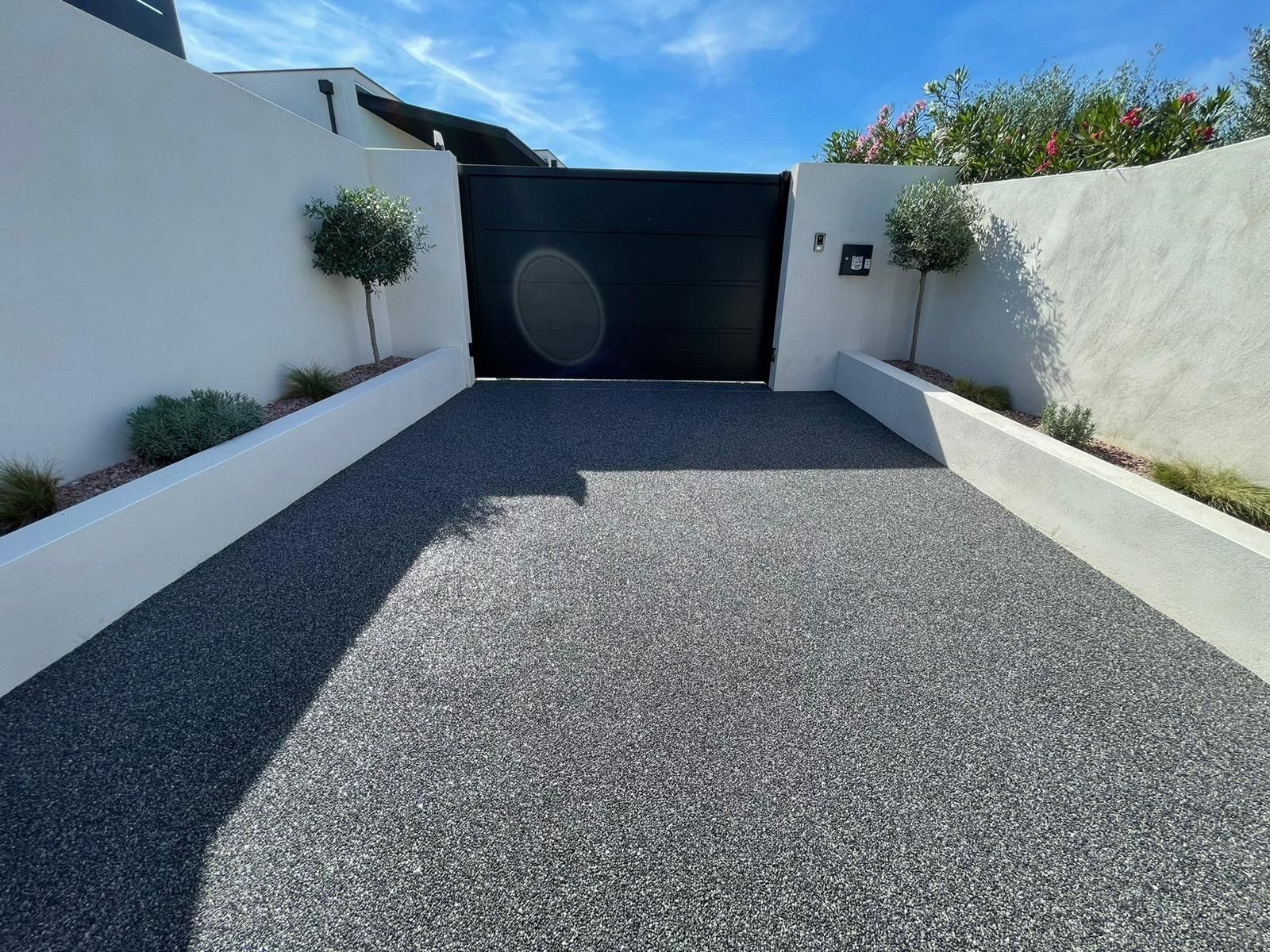 Allée en gravier noir avec des murs blancs et bord aménagé avec de la végétation.