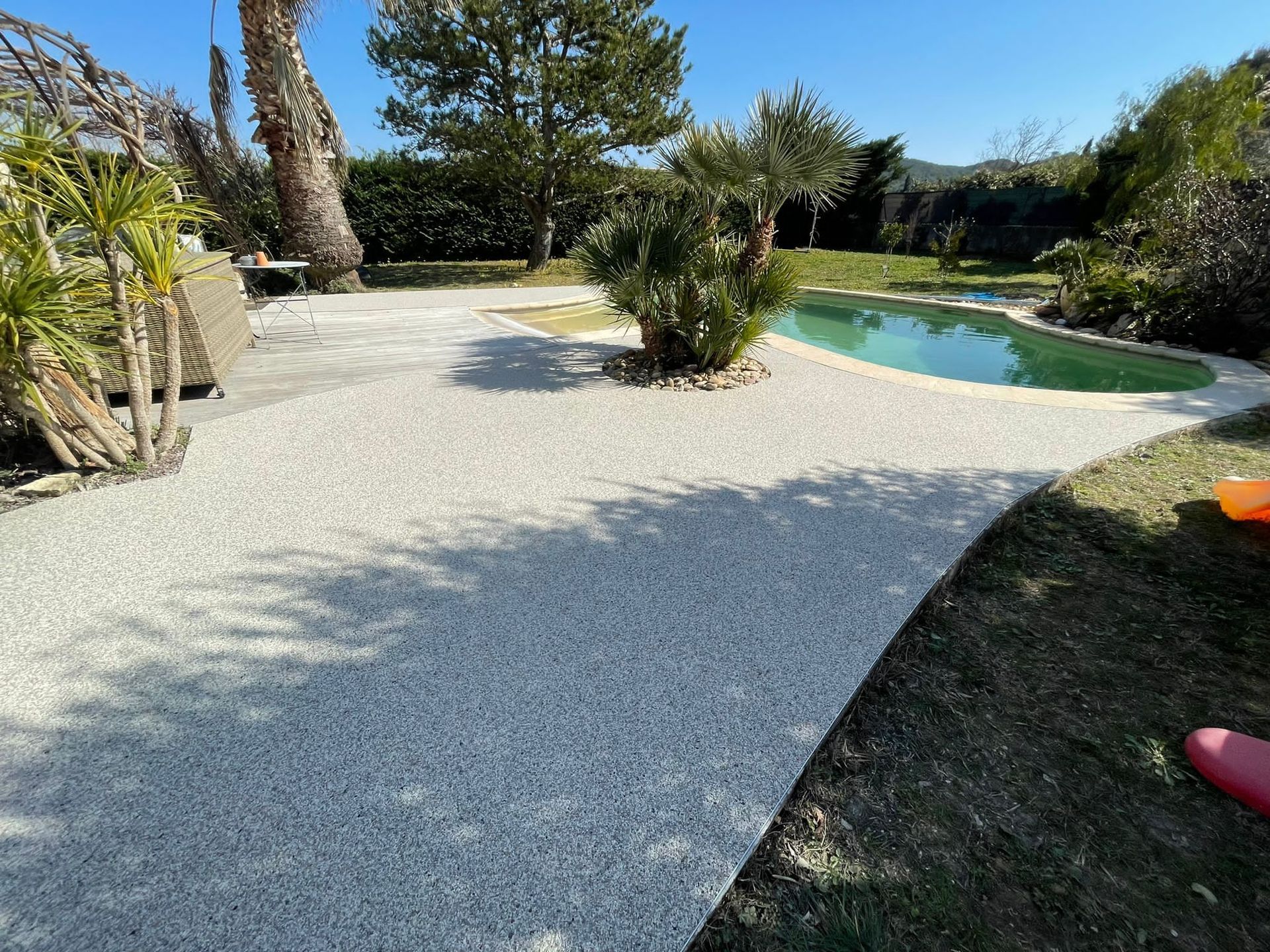 Piscine entourée de gravier blanc et de végétation, avec des palmiers et des arbres en arrière-plan.