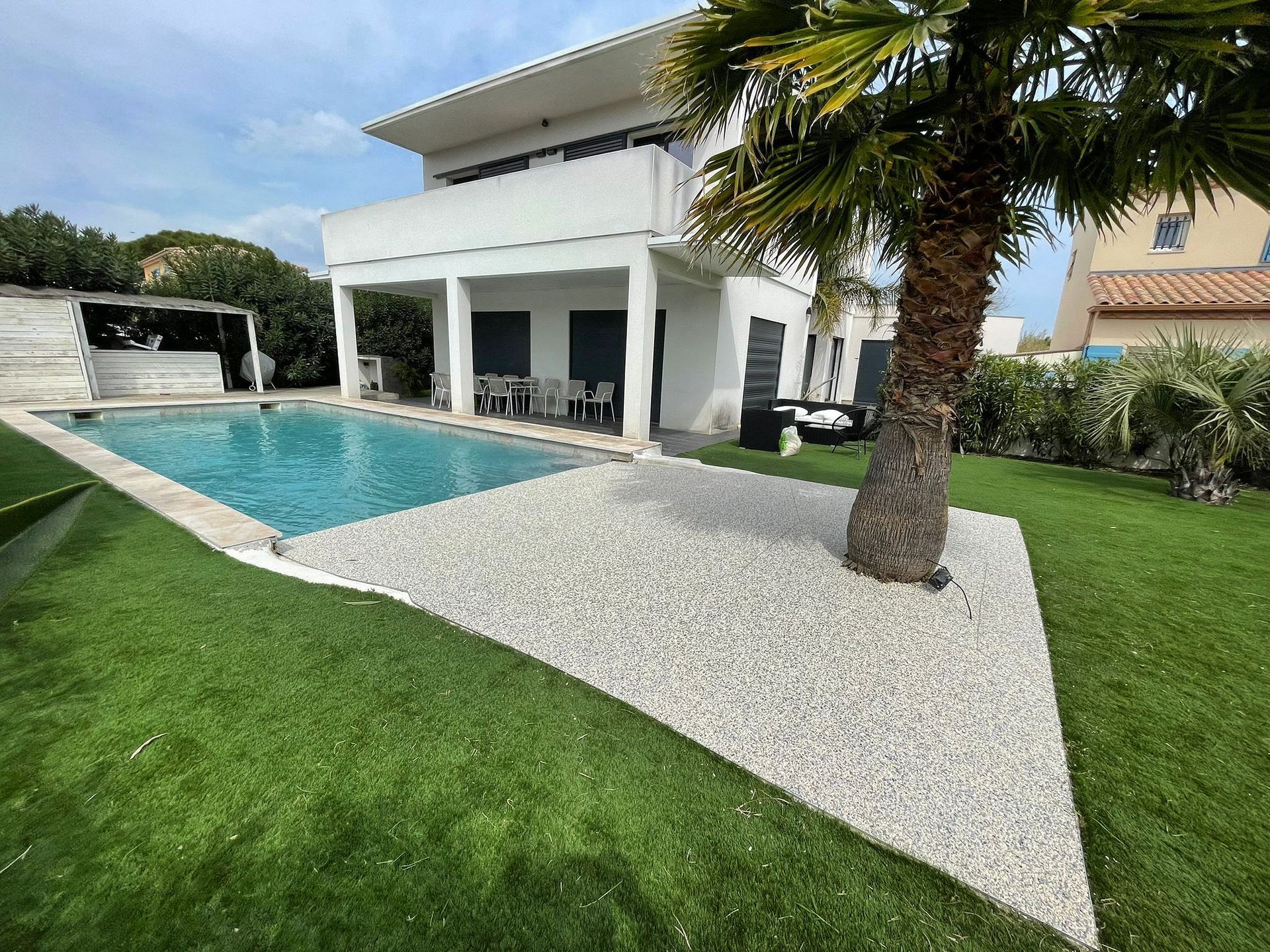 Maison moderne avec piscine et allée en gravier blanc, bordée de pelouse et de palmiers.