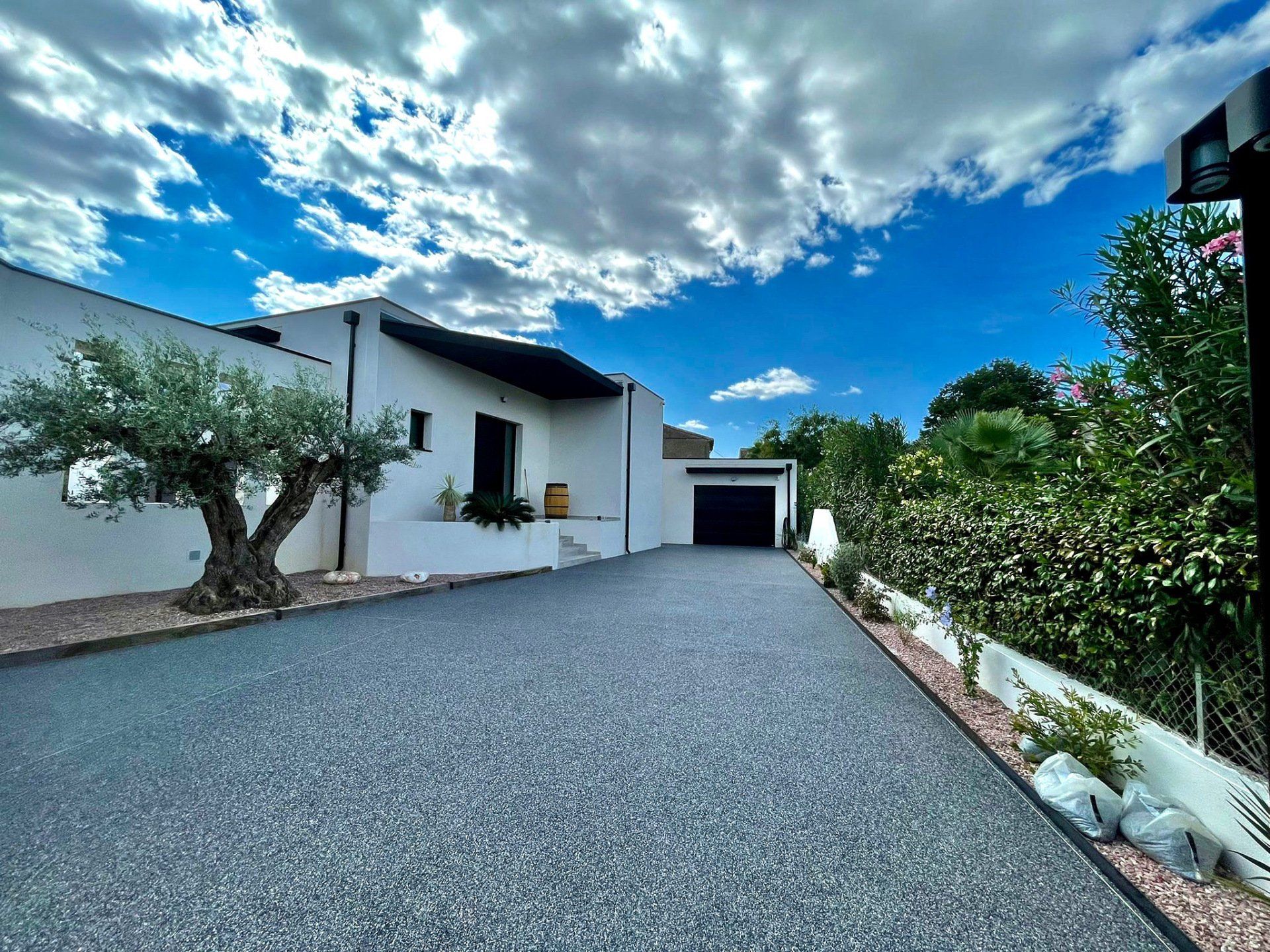 Allée en gravier gris menant à une maison blanche avec un arbre et un ciel bleu.