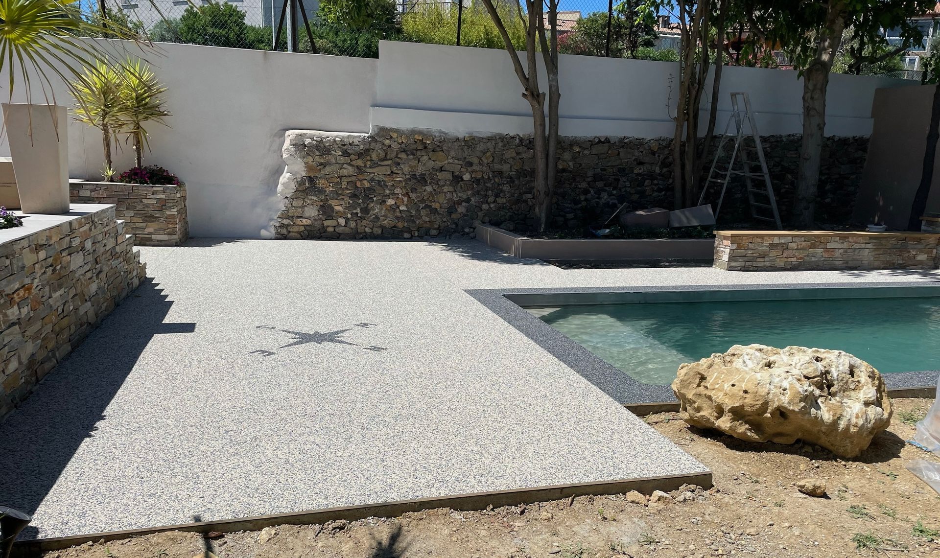 Piscine entourée de gravier blanc et de végétation, avec un mur en pierre.