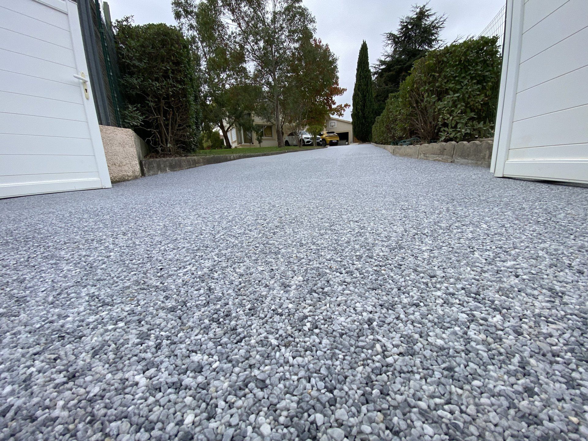 Zoom sur une allée en gravier gris avec portail blanc qui s'ouvre sur l'allée.