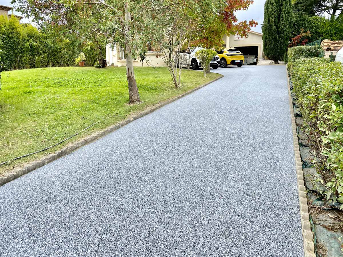 Allée en gravier gris bordée de pelouse verte et d'arbres.