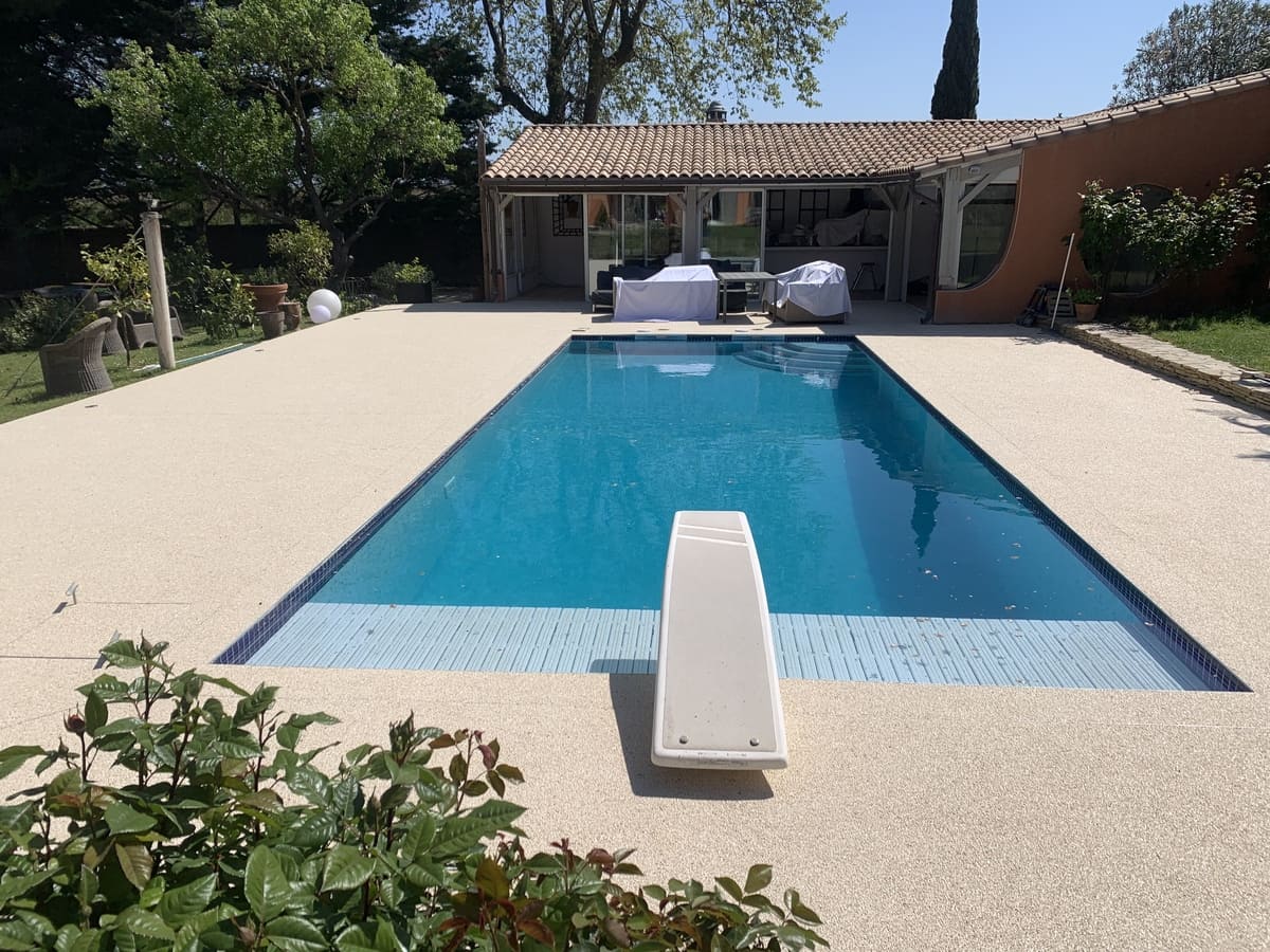 Piscine rectangulaire entourée de gravier beige avec un plongeoir et des arbres en arrière-plan.