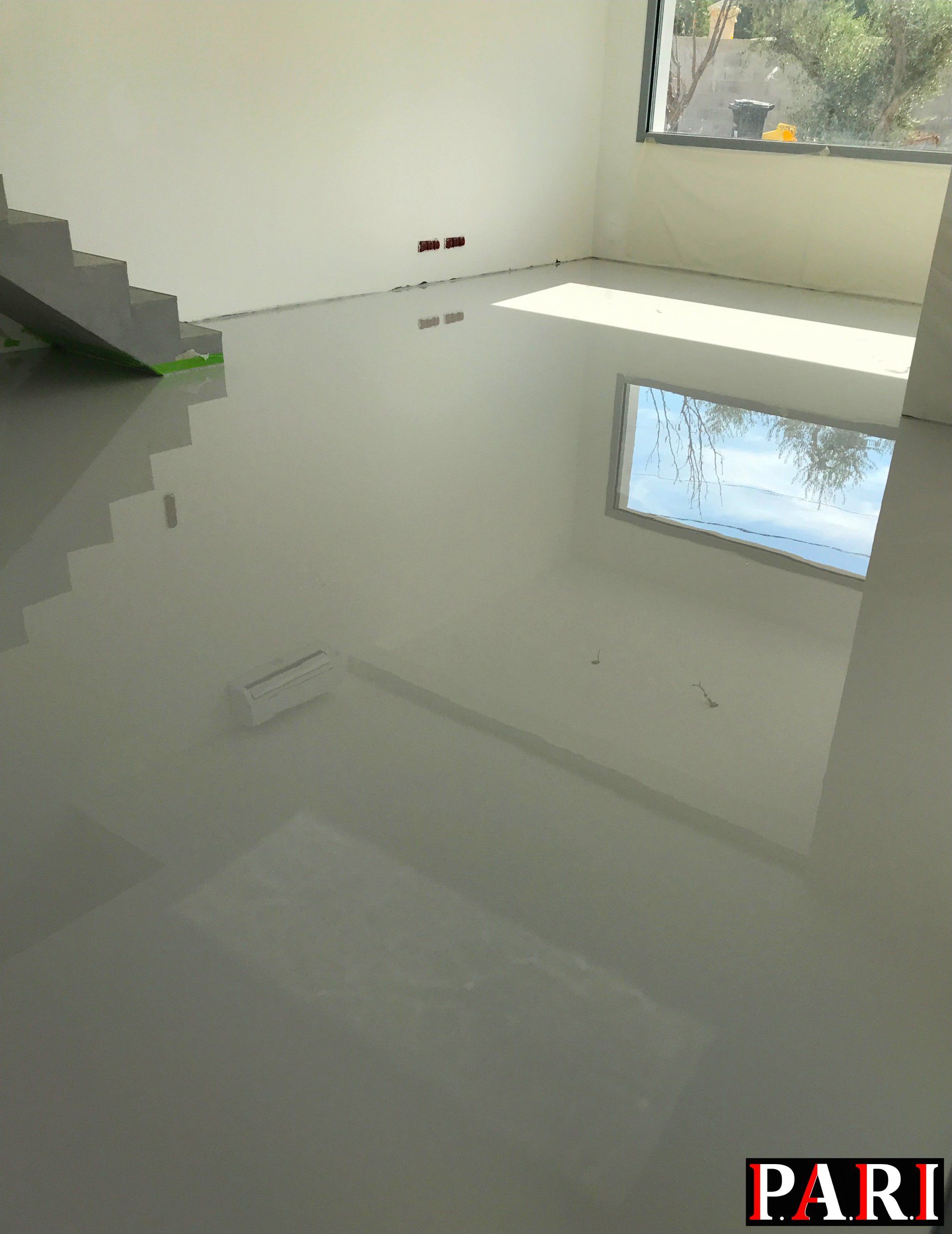 Zoom sur un sol en résine grise avec reflet de lumière sur le sol ou on peut voir une fenêtre et un escalier.