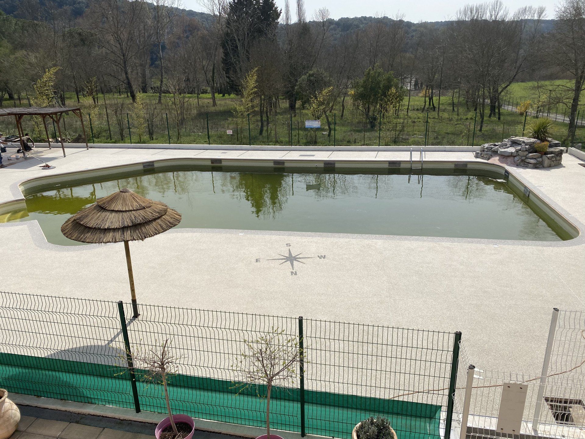 Piscine rectangulaire entourée de gravier blanc avec une clôture en fil de fer et des arbres en arrière-plan.