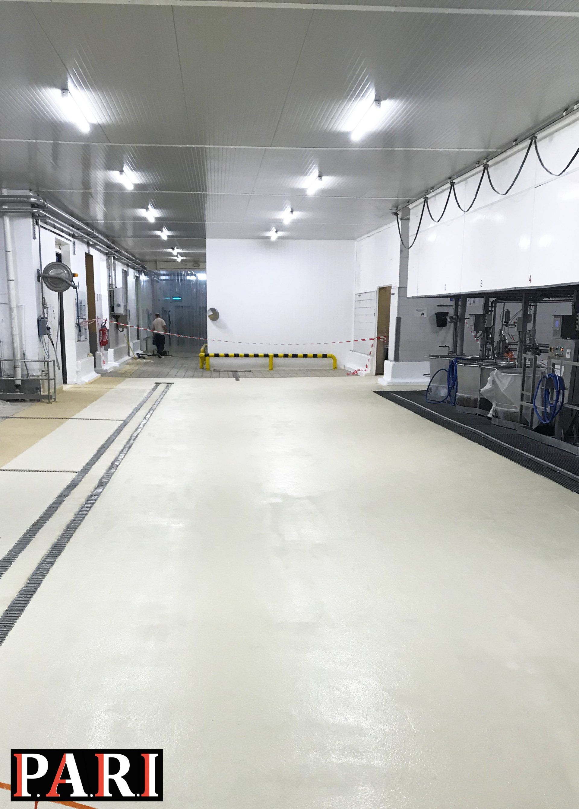 Sol en résine beige dans un grand entrepôt industriel avec des machines en arrière-plan.