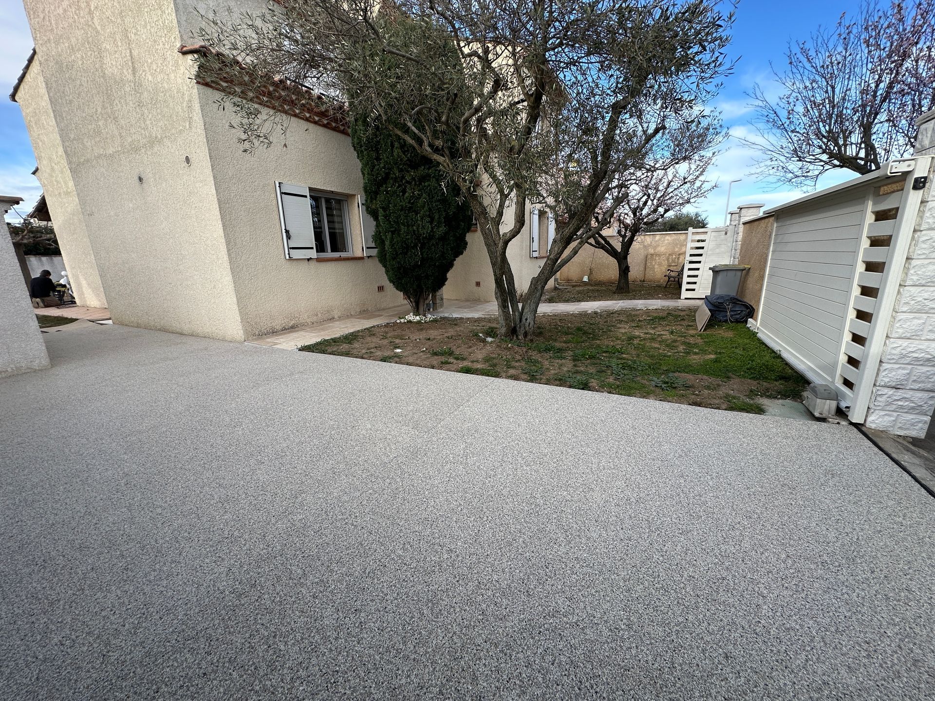 Allée extérieure en gravier gris devant une maison blanche avec des arbres.