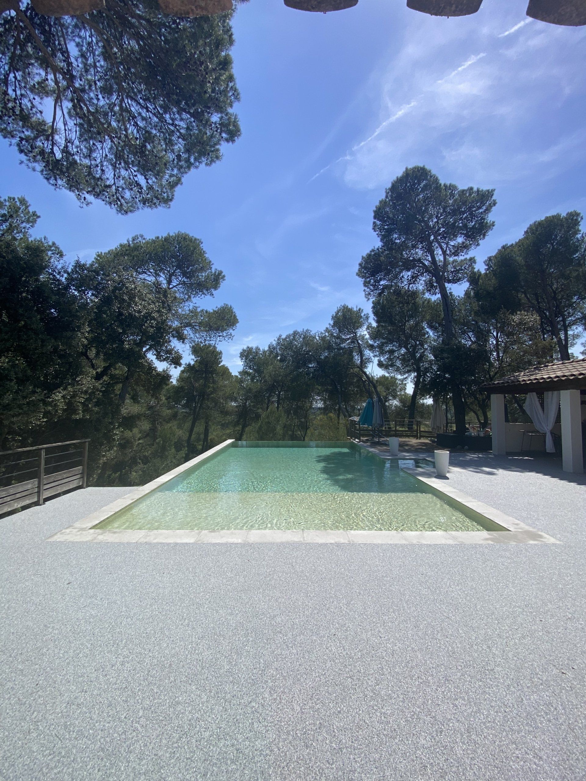 Piscine rectangulaire entourée de gravier beige avec des arbres et un ciel bleu.