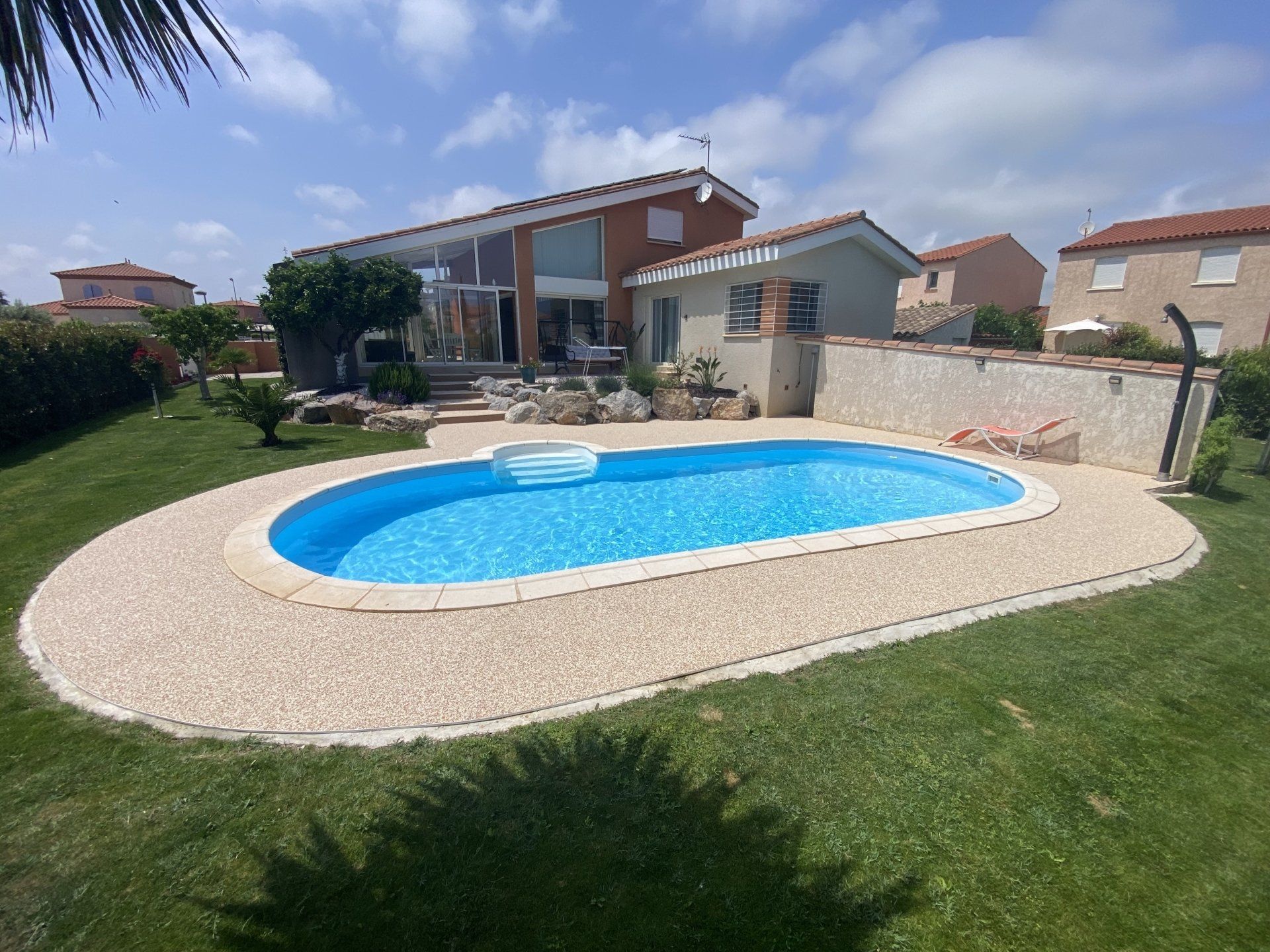 Piscine ovale entourée de gravier beige avec une pelouse verte et une maison en arrière-plan.