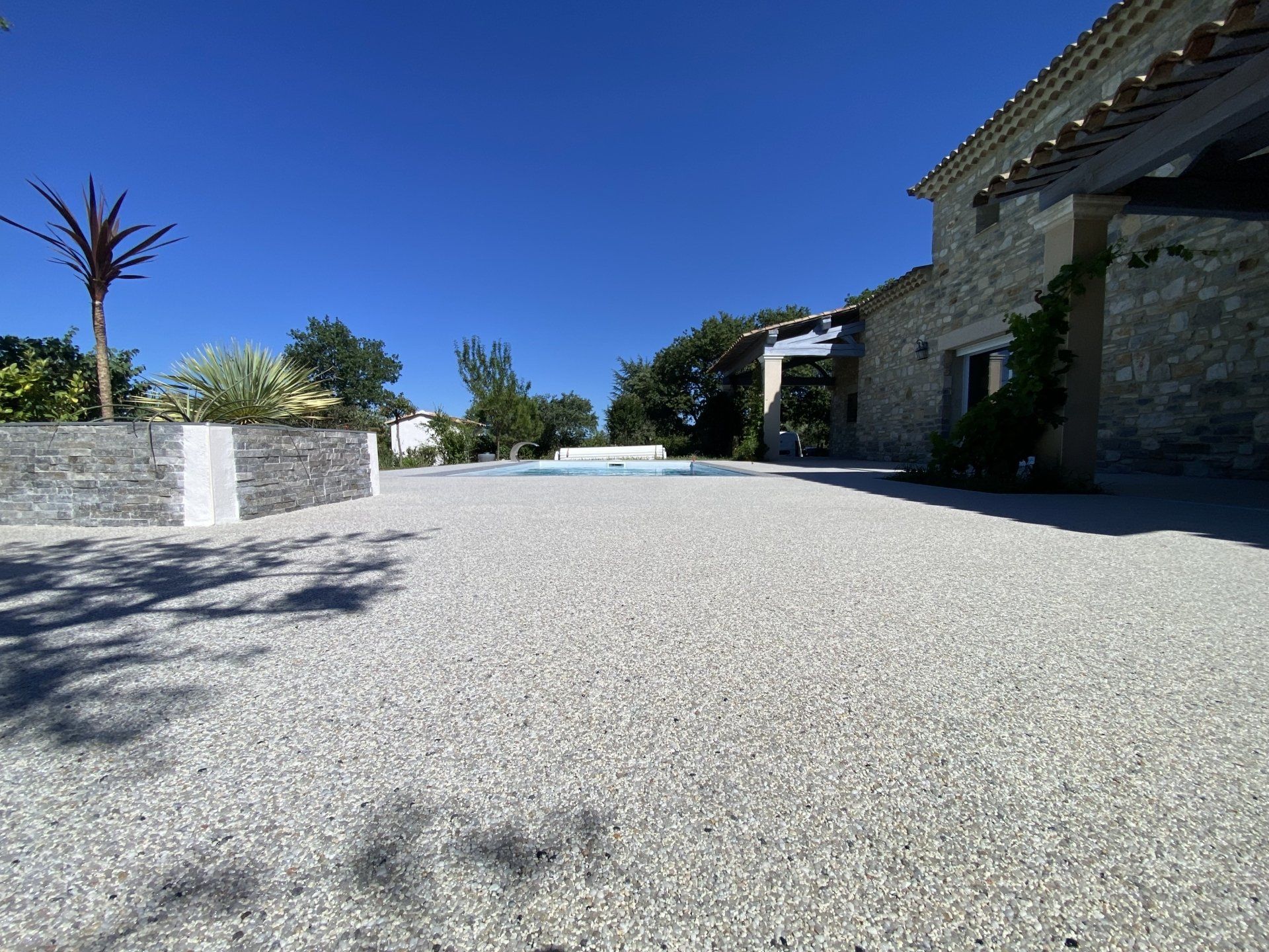 Allée en gravier beige menant à une maison en pierre avec des arbres et un ciel bleu.