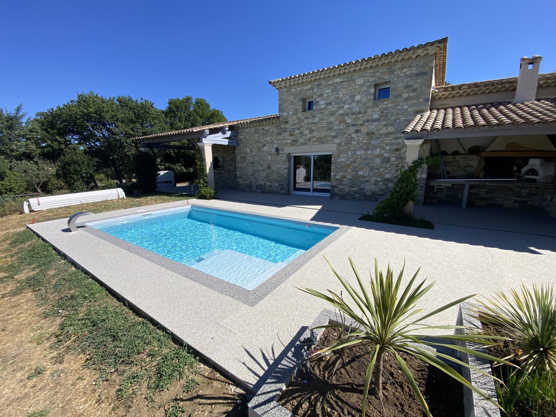Piscine entourée de gravier beige avec des plantes et une maison en pierre sous un ciel bleu.