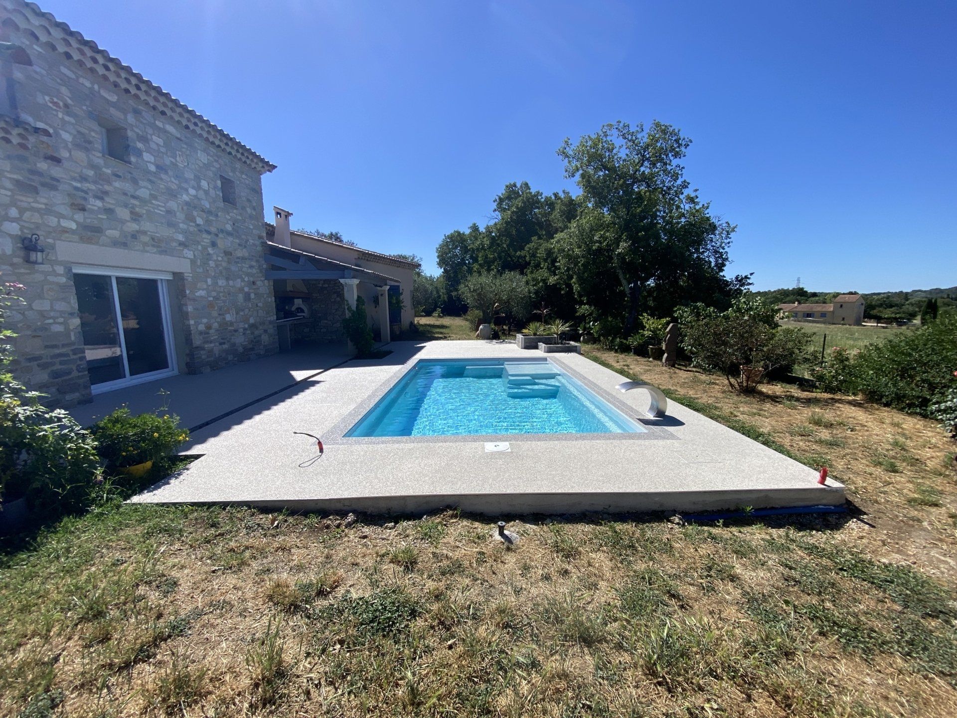 Piscine rectangulaire entourée de gravier beige, avec une maison en pierre en arrière-plan.