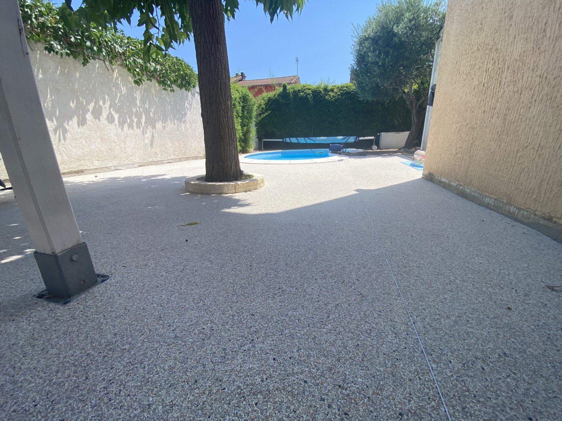 Allée en gravier beige menant à une piscine, avec un arbre et un mur en béton.