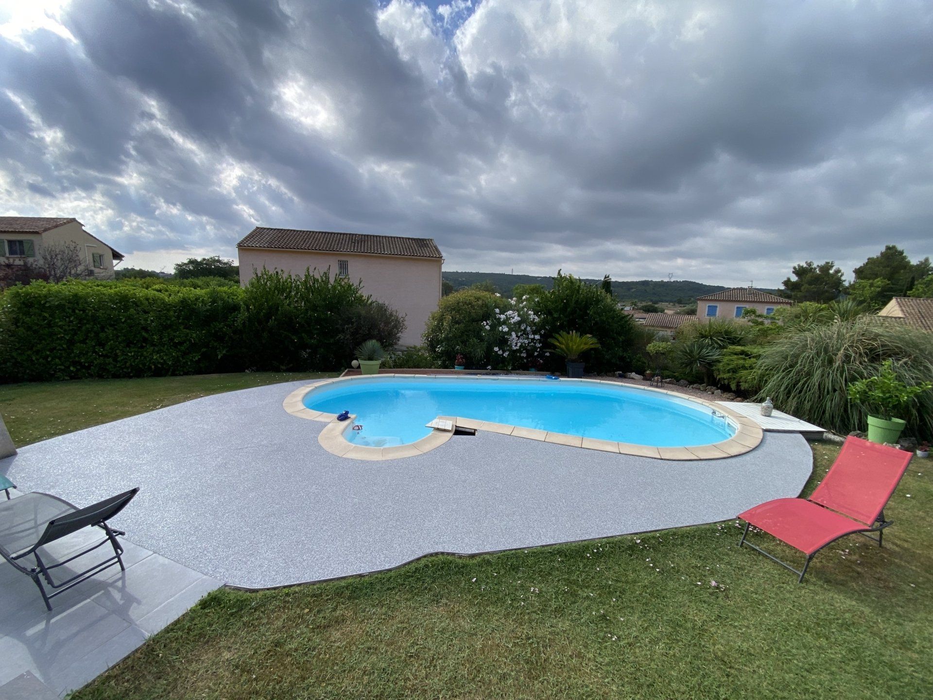Piscine entourée de gravier blanc avec des transats rouges et un jardin en arrière-plan.