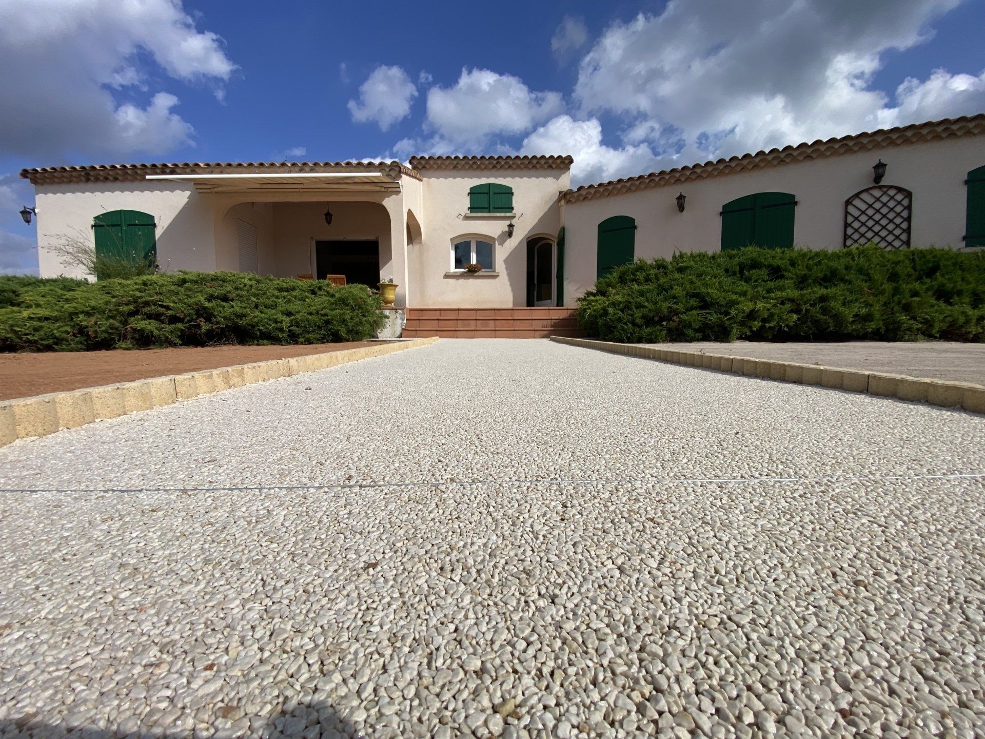 Allée en gravier blanc menant à une maison avec des volets verts et un jardin.