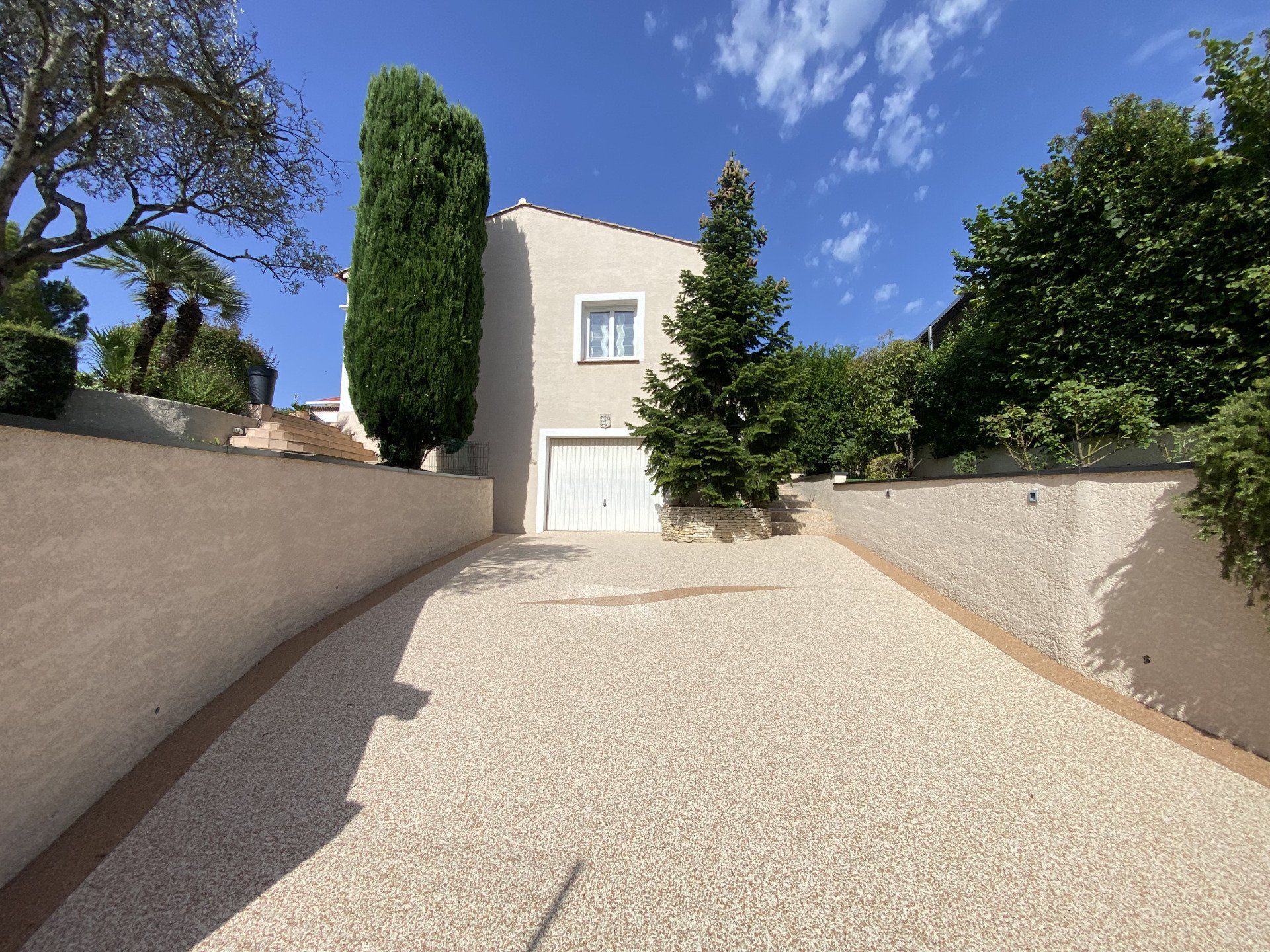 Allée en gravier blanc bordée de végétation et menant à une maison sous un ciel bleu.
