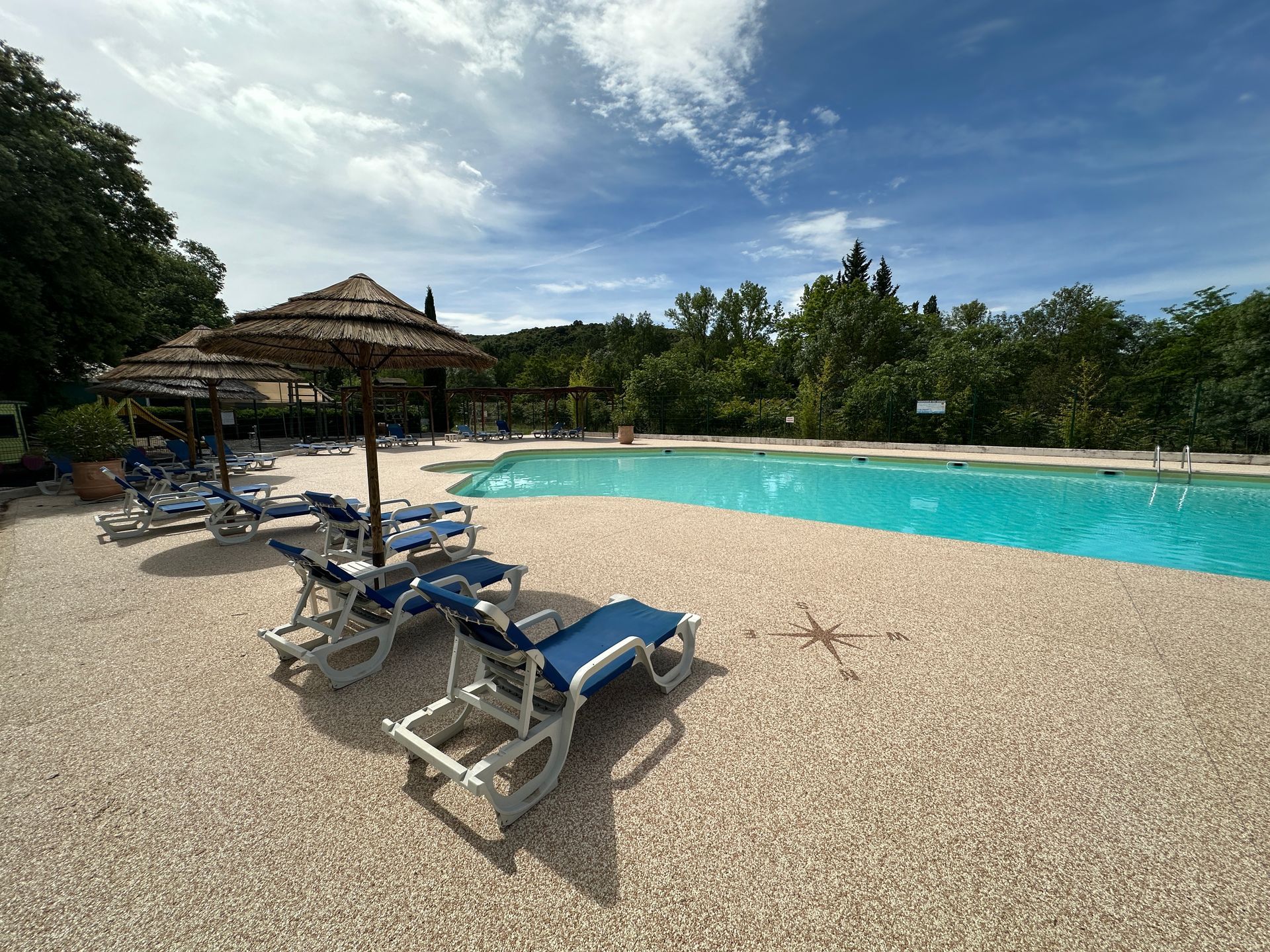 Plage de piscine en gravier avec transats et parasol, bordée de verdure.