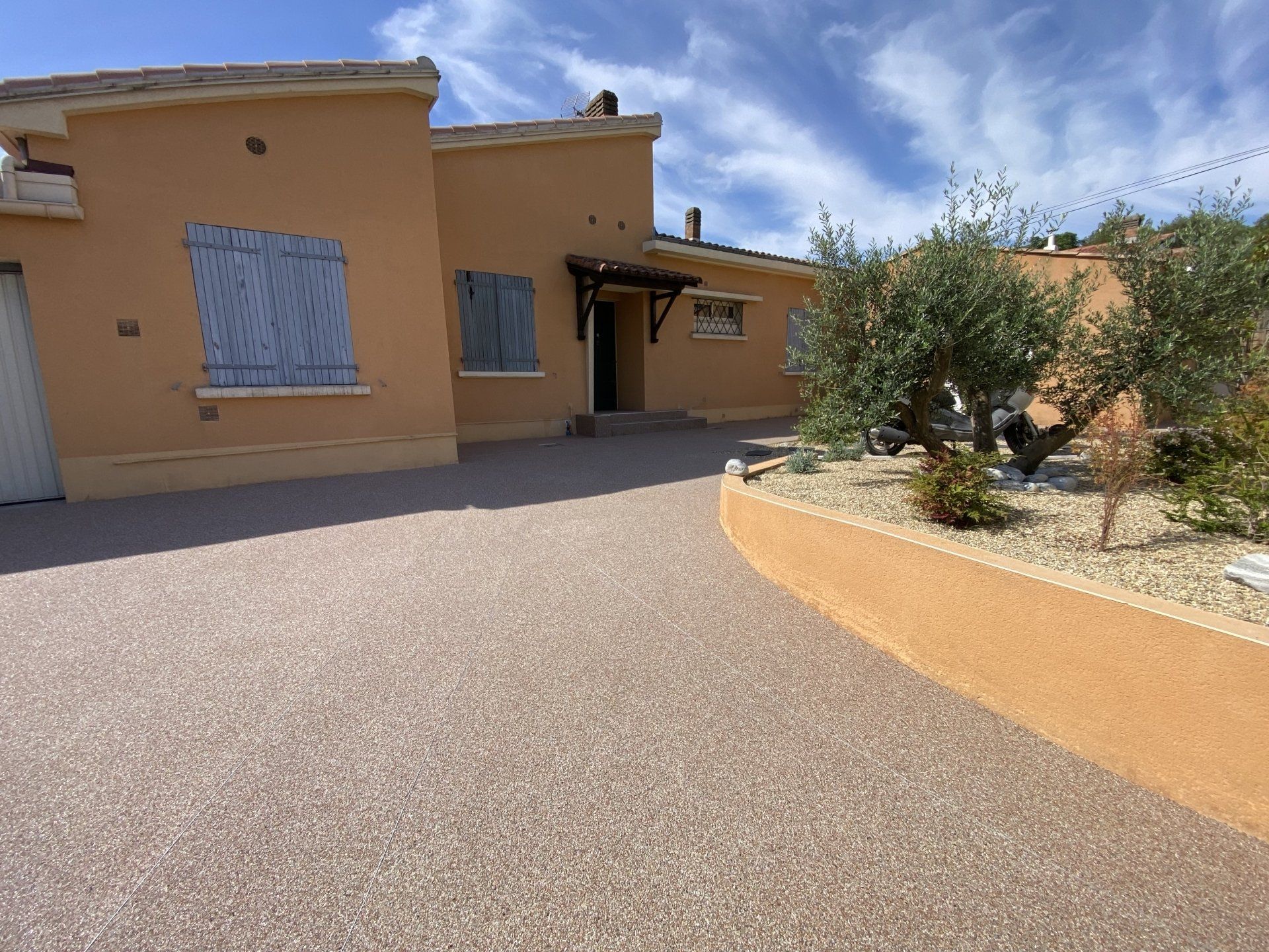 Allée en gravier rouge menant à une maison avec des volets bleus et un jardin.
