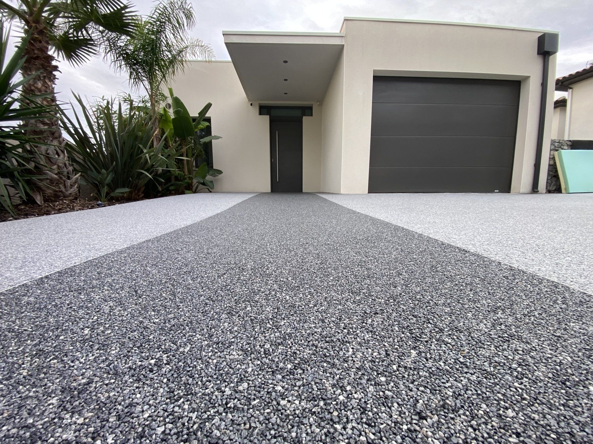 Allée en gravier gris menant à une maison avec un garage et des plantes en pot.