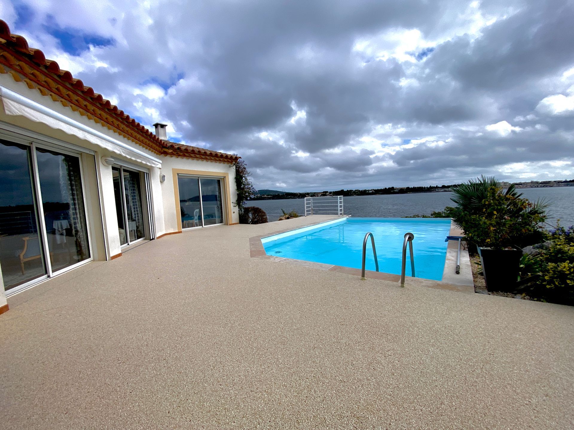 Piscine entourée de gravier blanc avec une maison en arrière-plan sous un ciel nuageux.