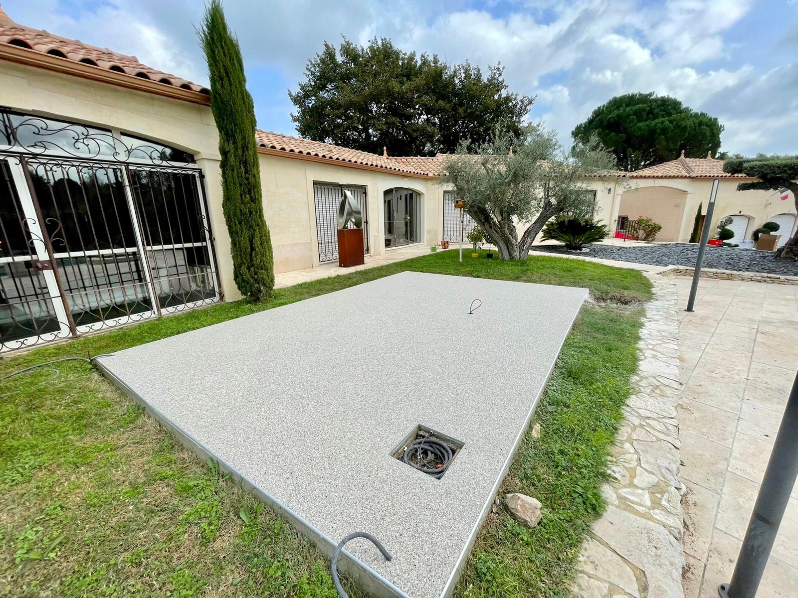 Allée en gravier blanc menant à une maison avec une protection baie vitrée en fer forgé et des arbres.