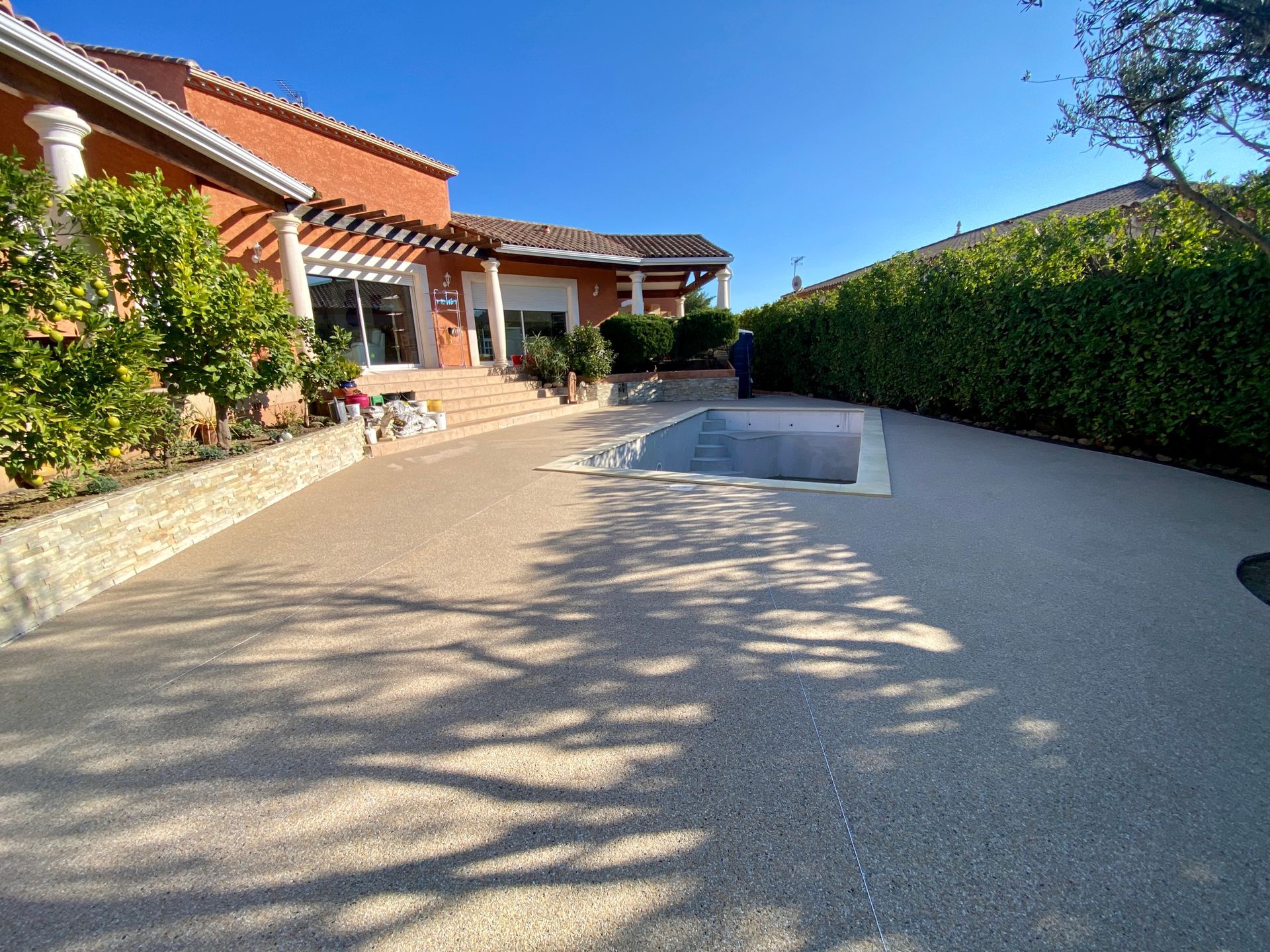 Allée en gravier menant à une maison rouge avec piscine et une haie verte et un ciel bleu.