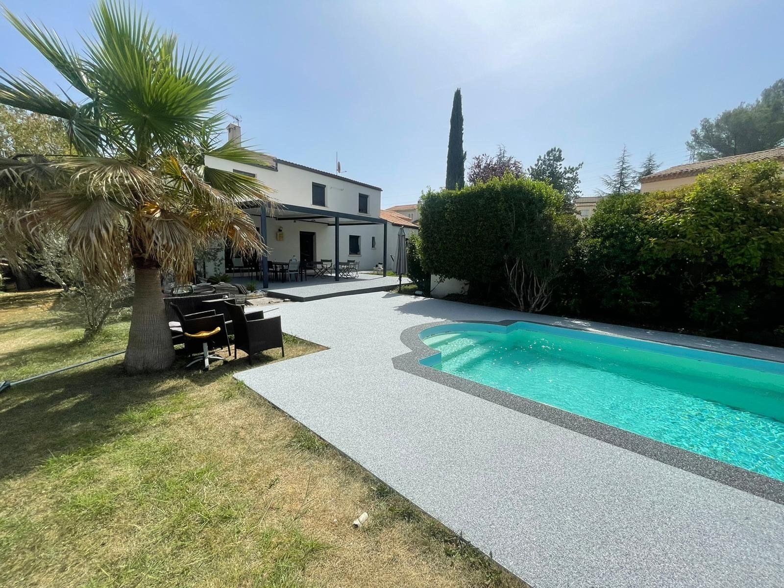 Allée en gravier beige menant à une piscine rectangulaire avec des arbres et une maison en arrière-plan.