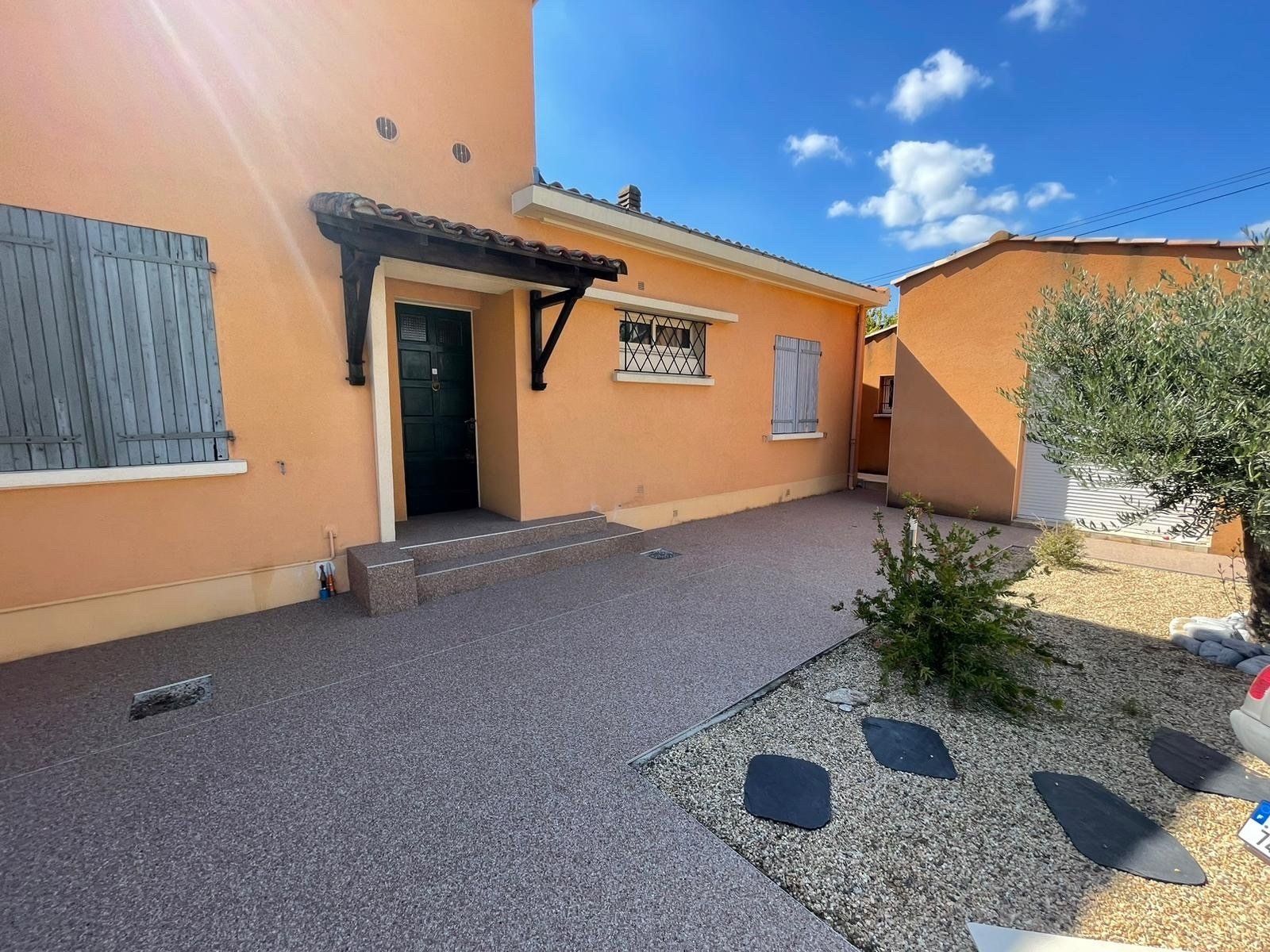 Façade d'une maison orange avec des volets bleus et une allée en gravier gris et massif végétal aménagé.