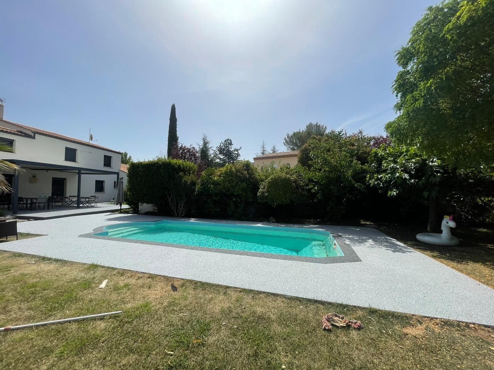 Piscine avec bord rond gris entouré de gravier gris avec une pelouse verte et une maison en arrière-plan.