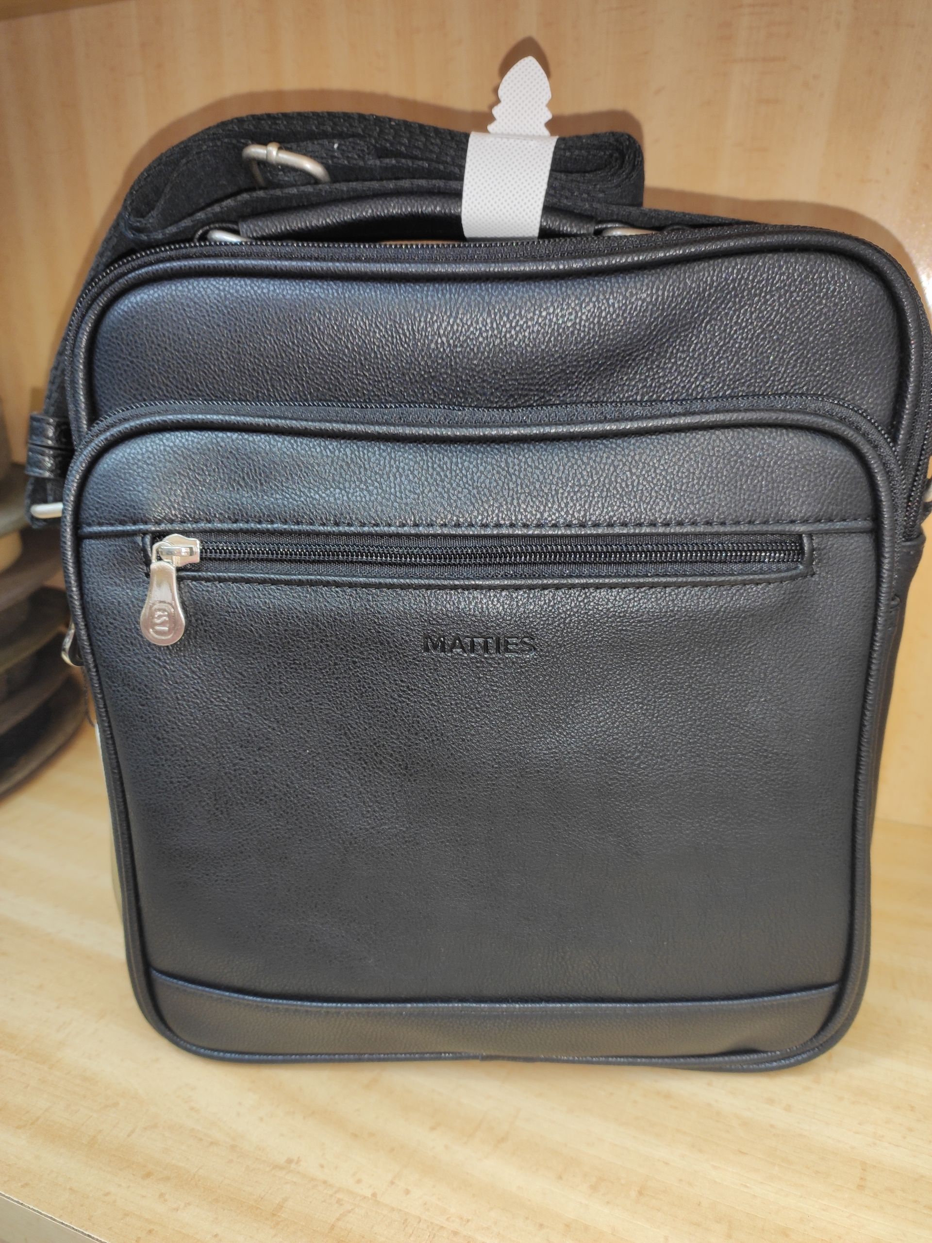 Un bolso mensajero de cuero negro con correa está sobre una mesa de madera.