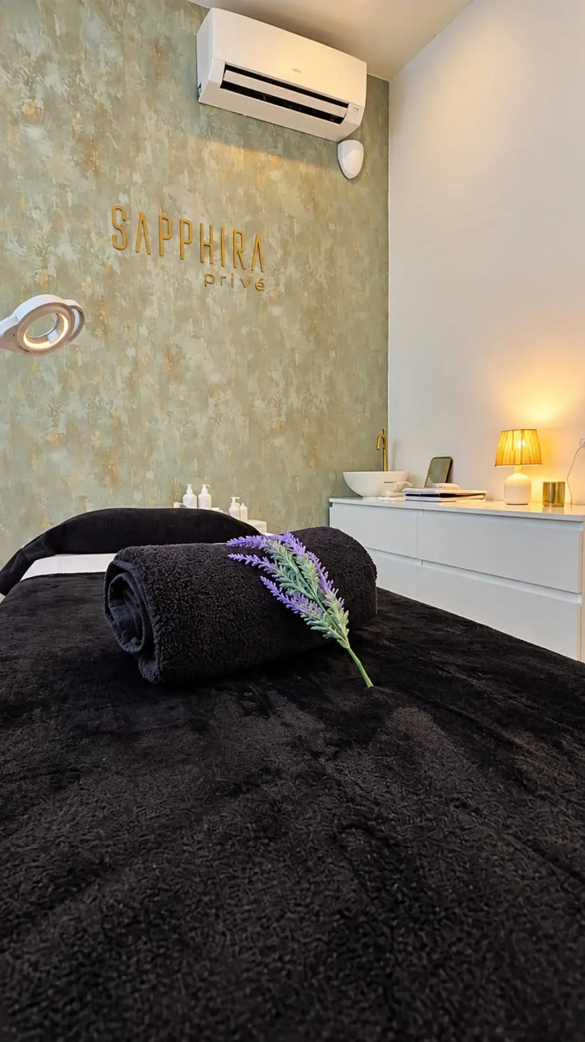 Massage room with a black treatment bed, rolled towel, wall art, and a wall-mounted air conditioner.