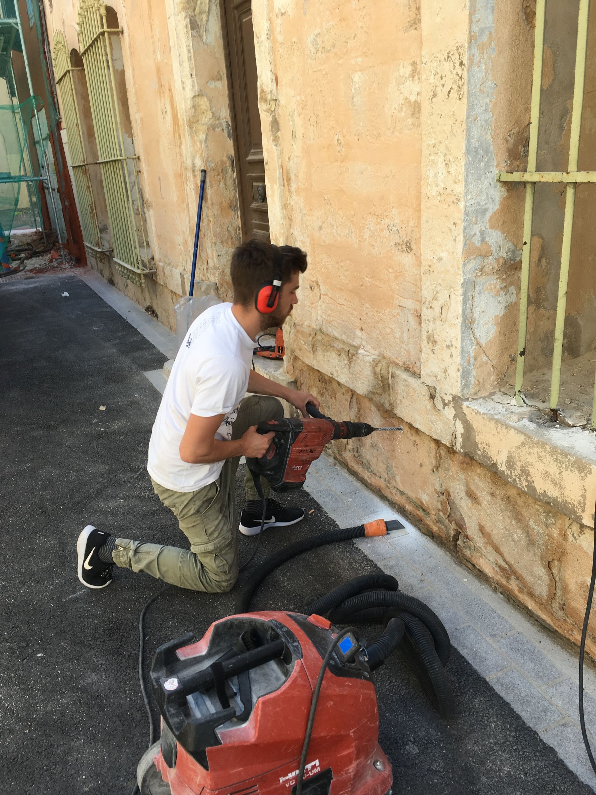 Un homme en train de percer un mur en pierre avec une perceuse électrique, vue de côté.