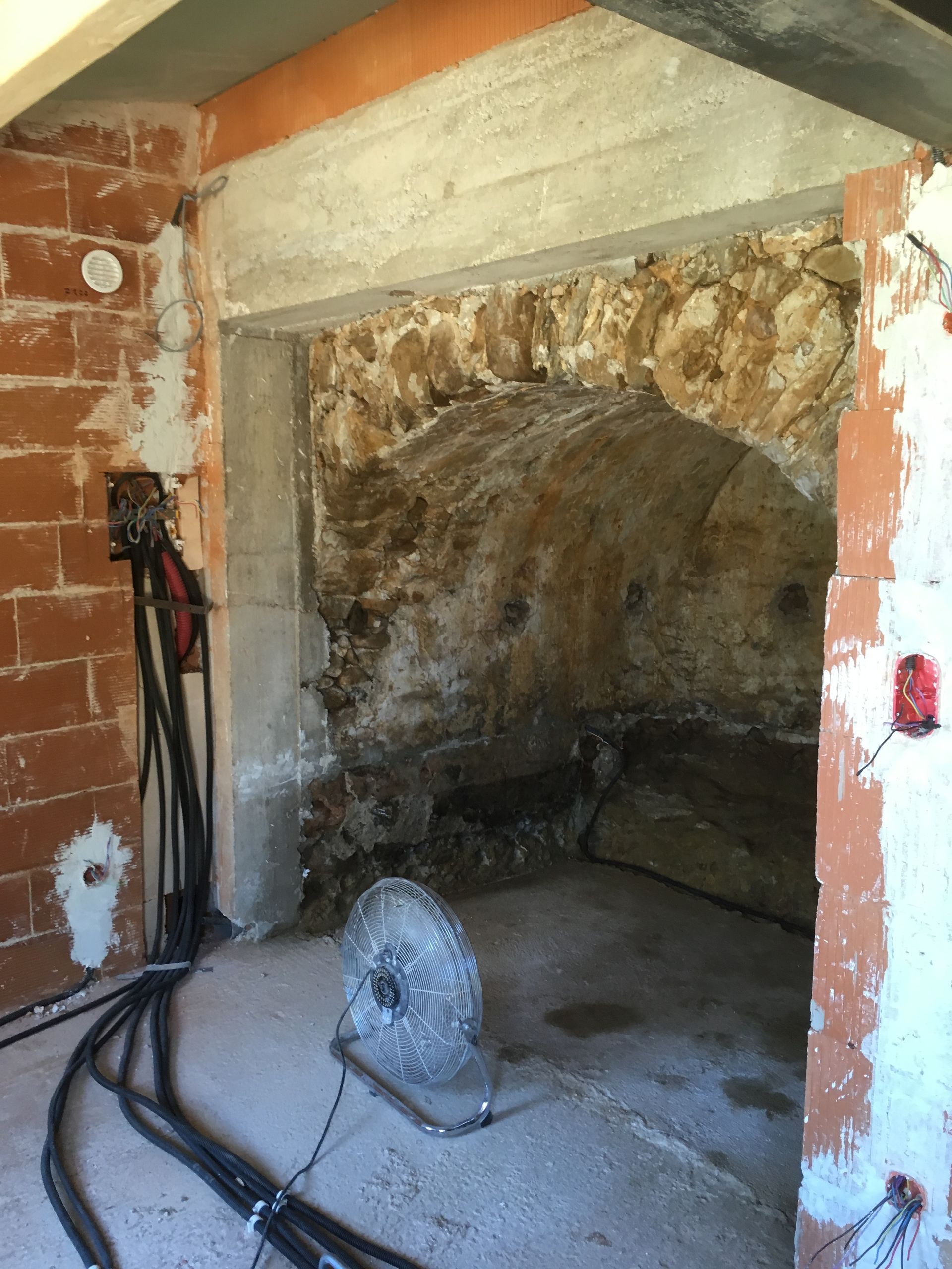 Vue d'un mur intérieur en pierre avec un ventilateur posé au sol.