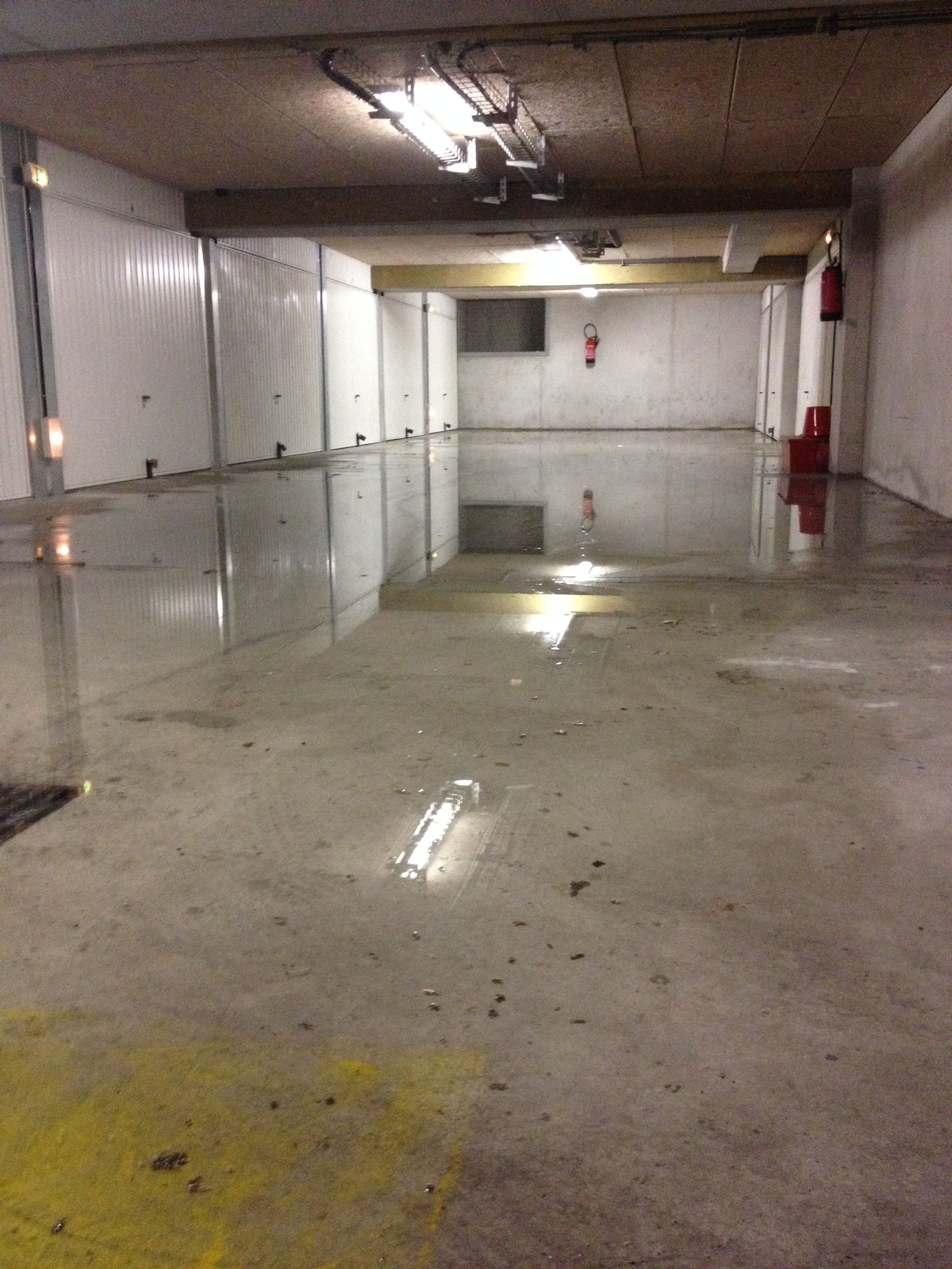 Un couloir souterrain ou un tunnel avec des murs en béton brut. Il y a des tuyaux et des câbles visibles le long des murs, ainsi que des lumières au plafond.