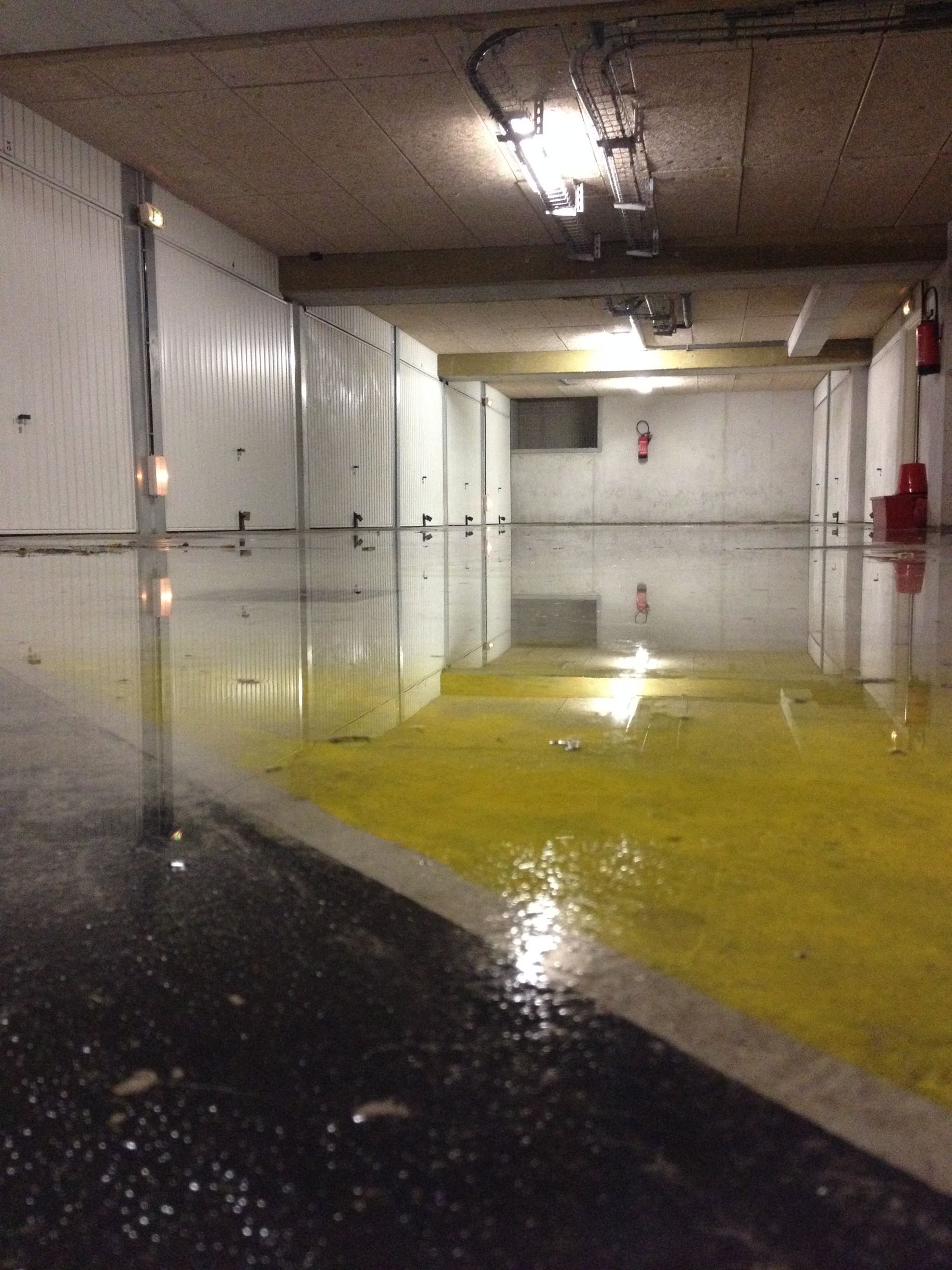 Un couloir souterrain ou un tunnel avec des murs en béton brut. Il y a des tuyaux et des câbles visibles le long des murs, ainsi que des lumières au plafond  montrant plus de détails du couloir et des équipements.