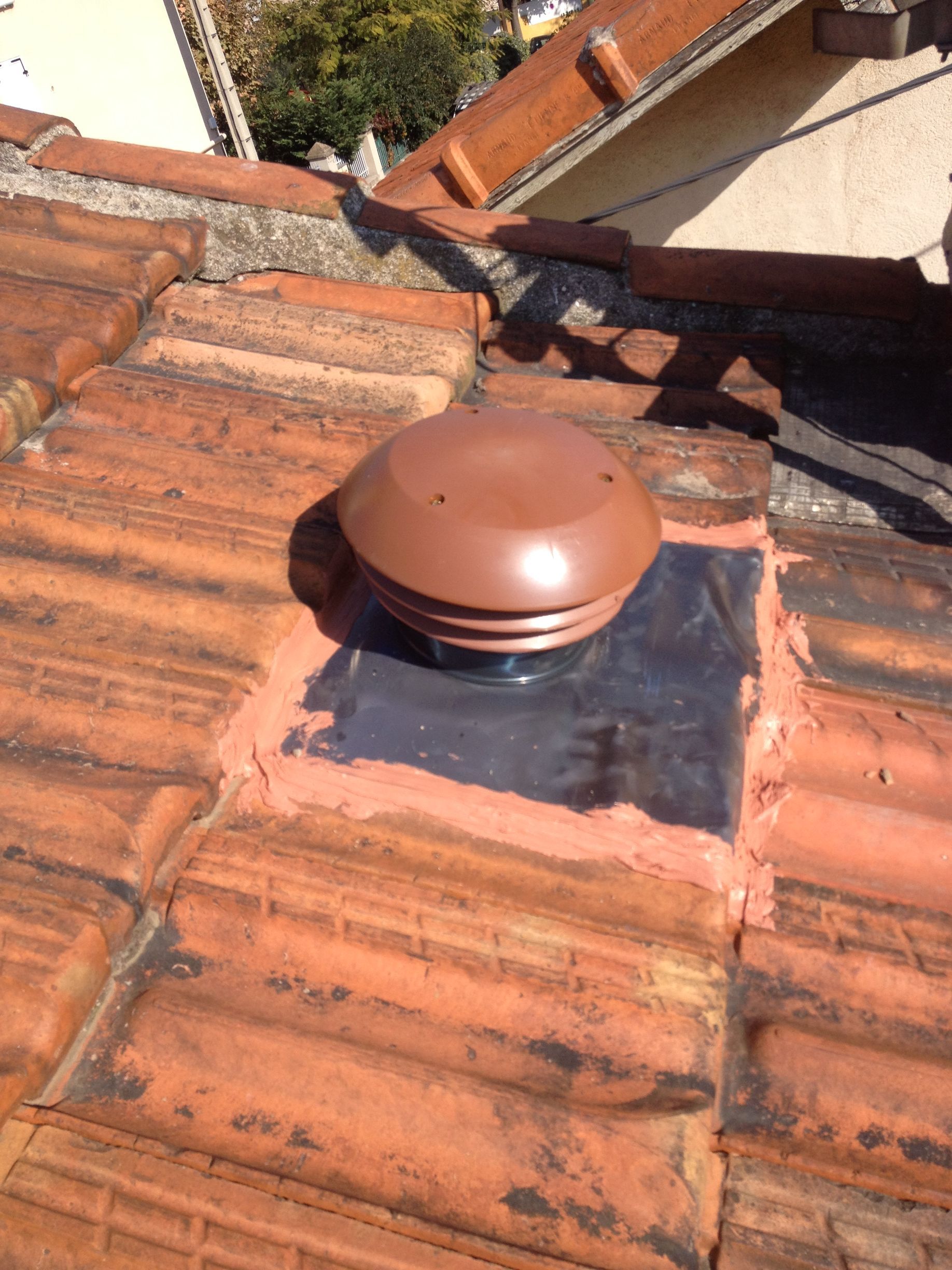 Un toit en tuiles avec une sortie de ventilation en plastique brun. La sortie est installée sur une plaque en métal noir, et les tuiles autour sont légèrement déplacées.