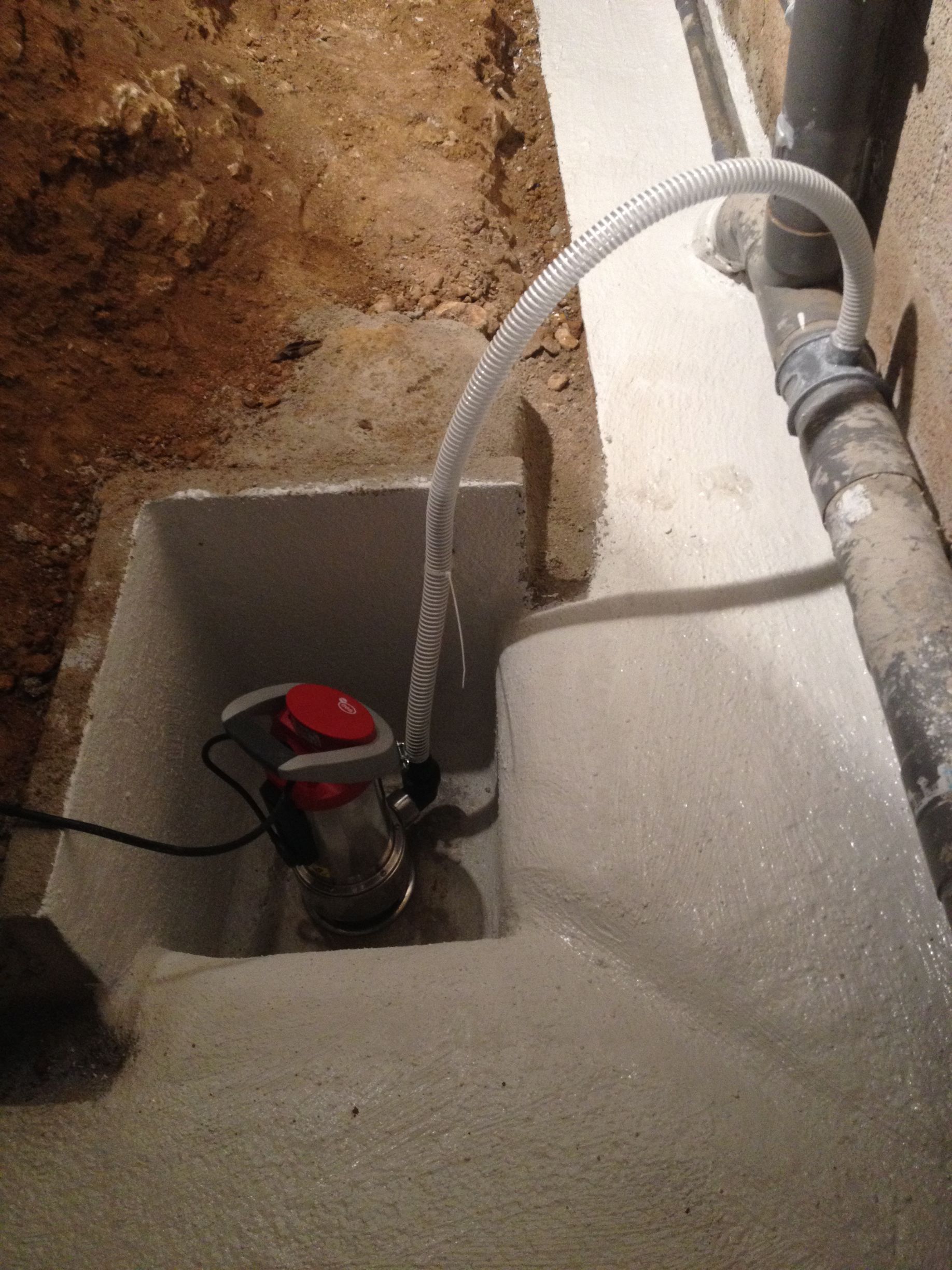 Un gros plan du dispositif électronique rouge et blanc fixé sur un tuyau en plastique blanc. Le dispositif semble être un capteur ou un détecteur dans une fosse.