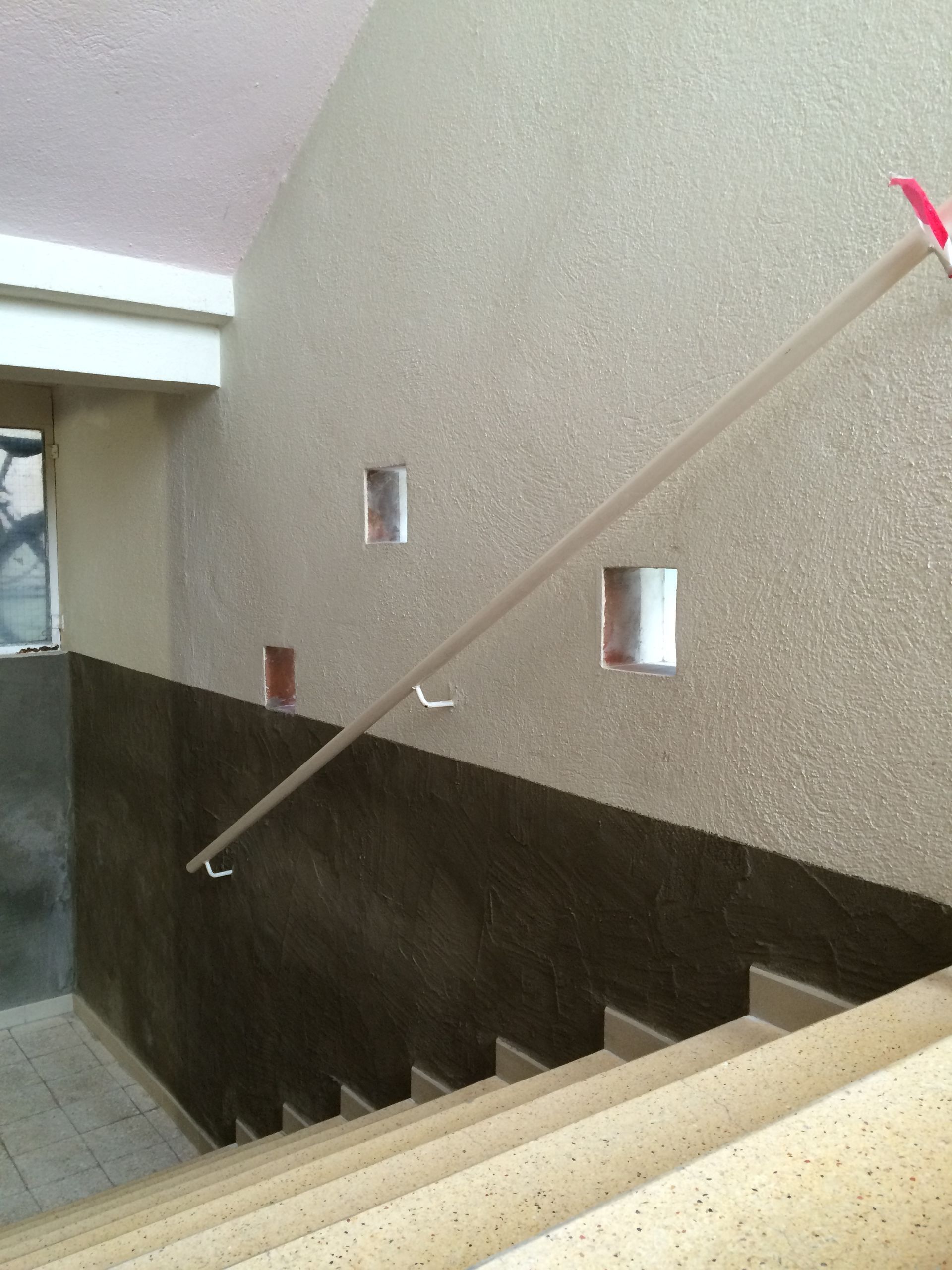 Escalier intérieur avec une rampe en bois clair. Le mur adjacent est partiellement recouvert d'un enduit gris texturé et beige lisse.