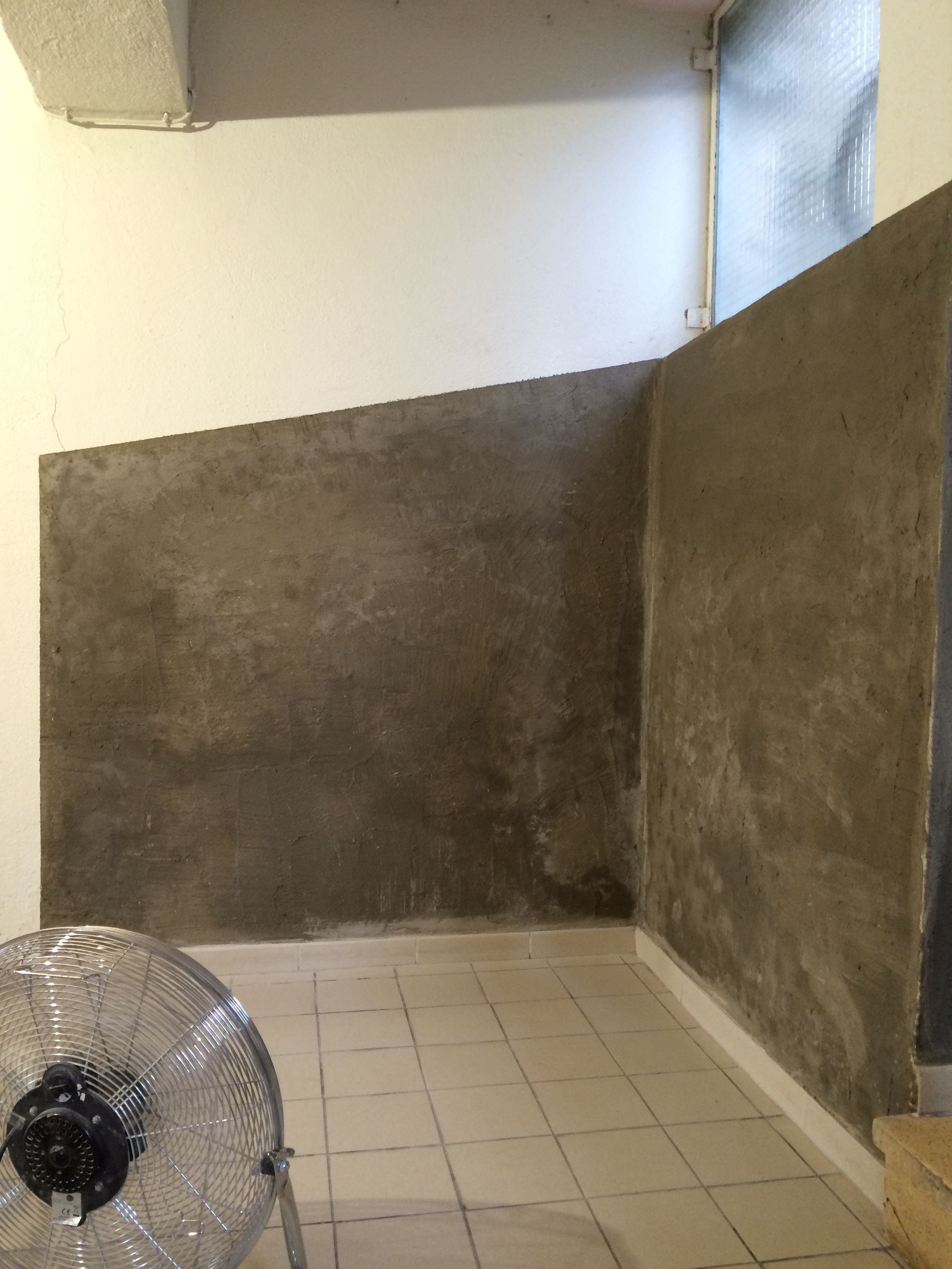 Mur intérieur en cours de rénovation avec une couche de plâtre gris appliquée. Un ventilateur est posé au sol.