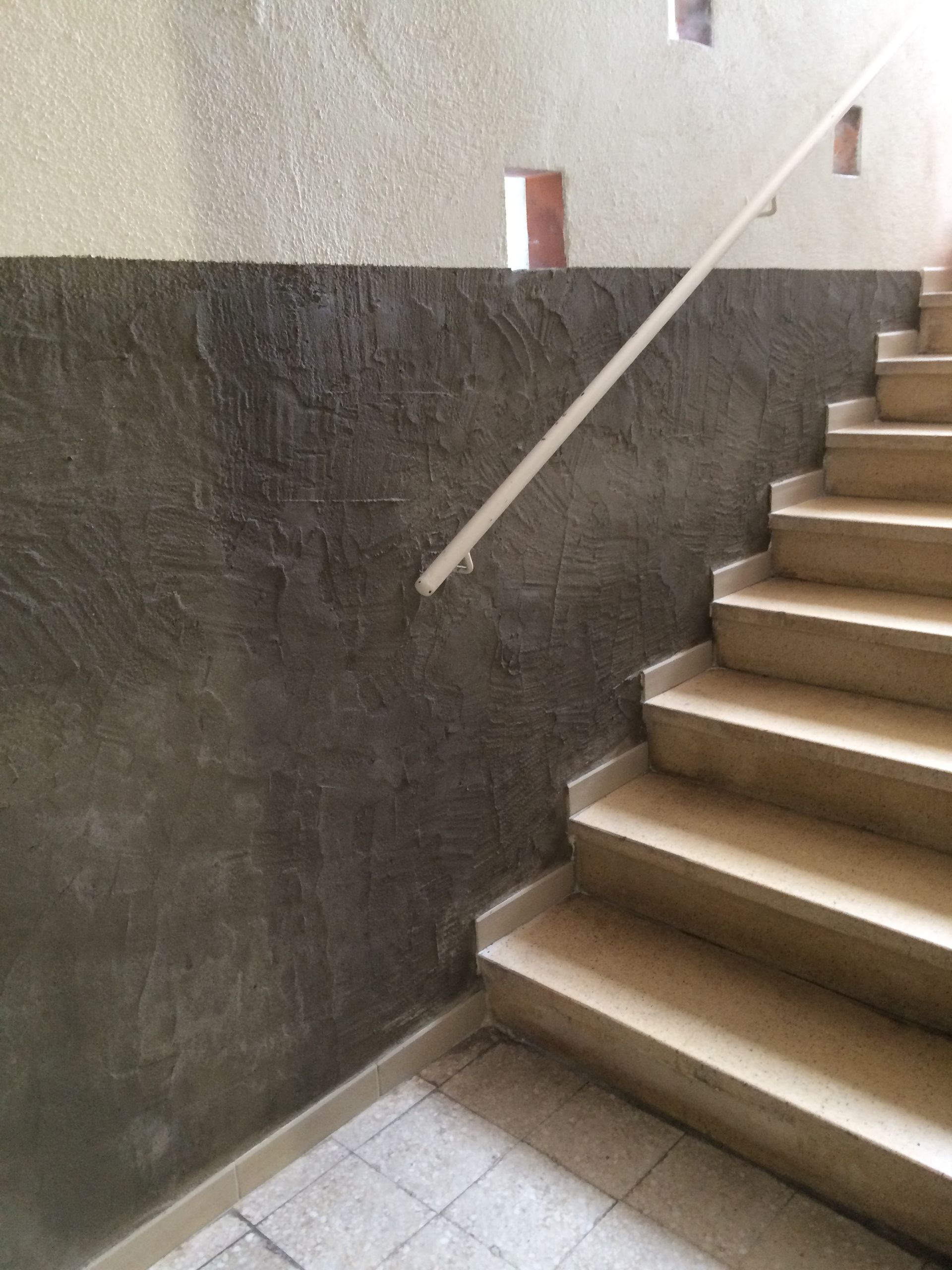 Escalier intérieur avec une rampe en bois clair. Le mur adjacent est recouvert d'un enduit gris texturé.