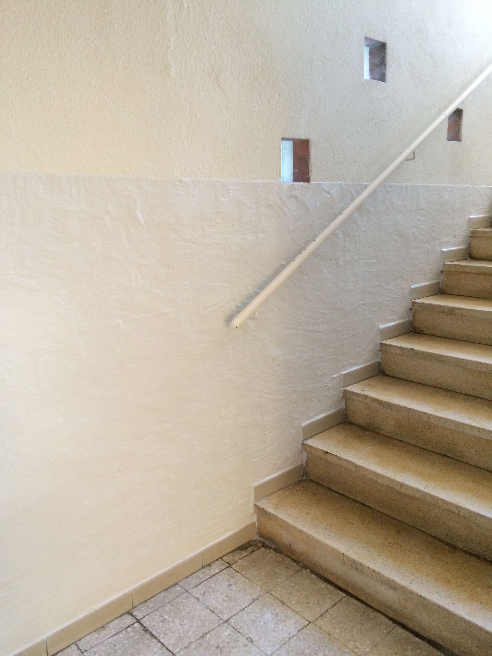 Escalier intérieur avec une rampe en bois clair. Le mur adjacent est recouvert d'un enduit beige lisse.